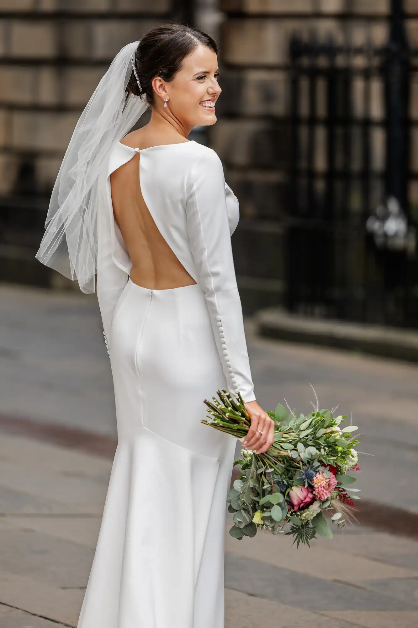 A smiling bride in a long-sleeved white satin gown with an open back stands outdoors, holding a bouquet of flowers with greenery and pink blooms. She has a veil in her hair and faces slightly away, displaying the dress’s button details and elegant silhouette.