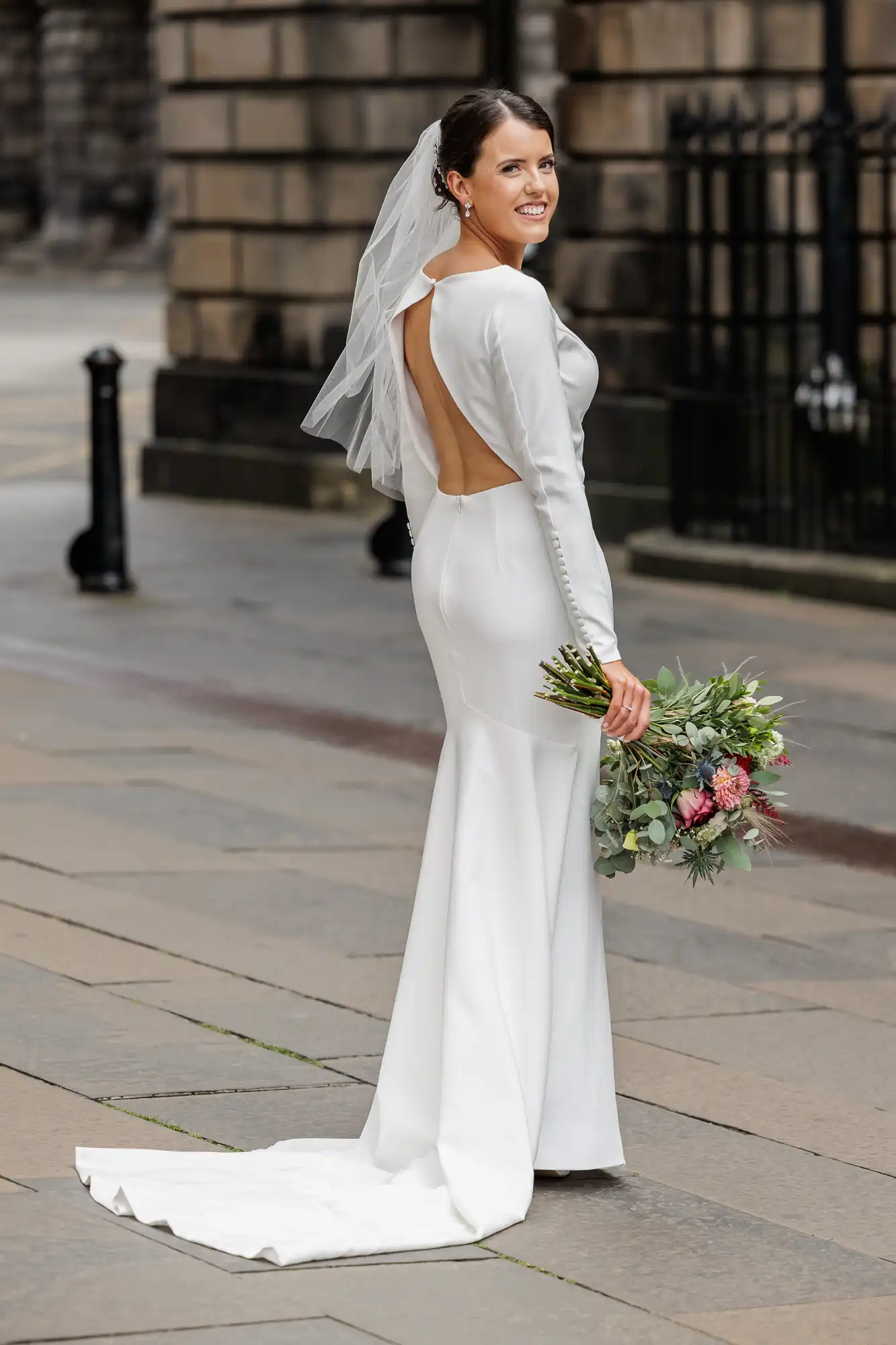 A bride in a sleek, long-sleeved white dress with an open back and train stands outdoors on stone pavement, smiling over her shoulder. She wears a veil and holds a bouquet of pink and green flowers. Stone buildings and black railings are in the background.