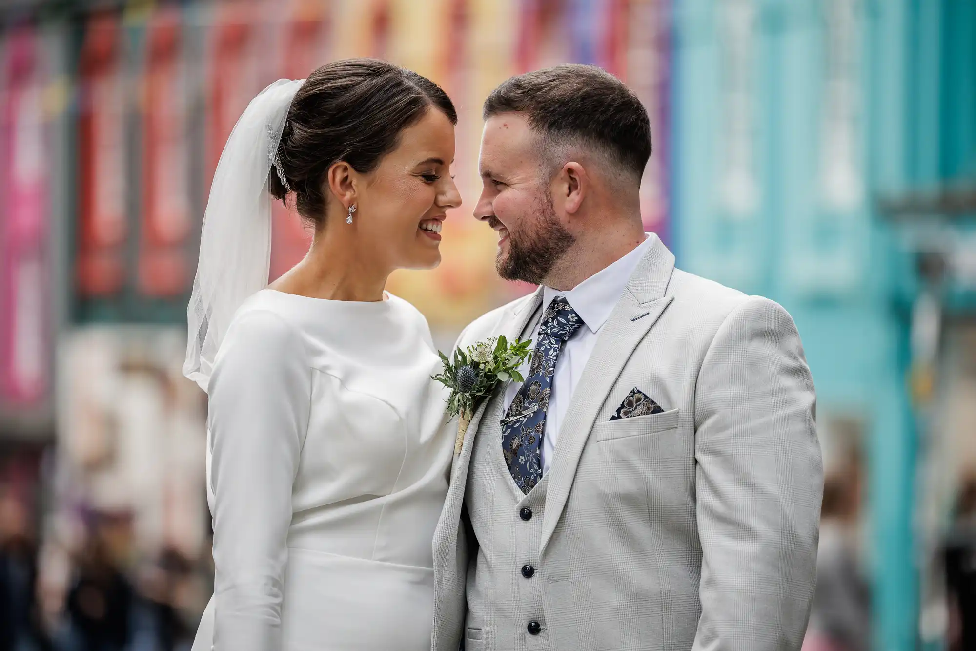 A smiling bride in a white dress and veil stands close to a groom in a light grey suit and floral tie. They gaze at each other warmly, touching foreheads, with colourful buildings blurred in the background.