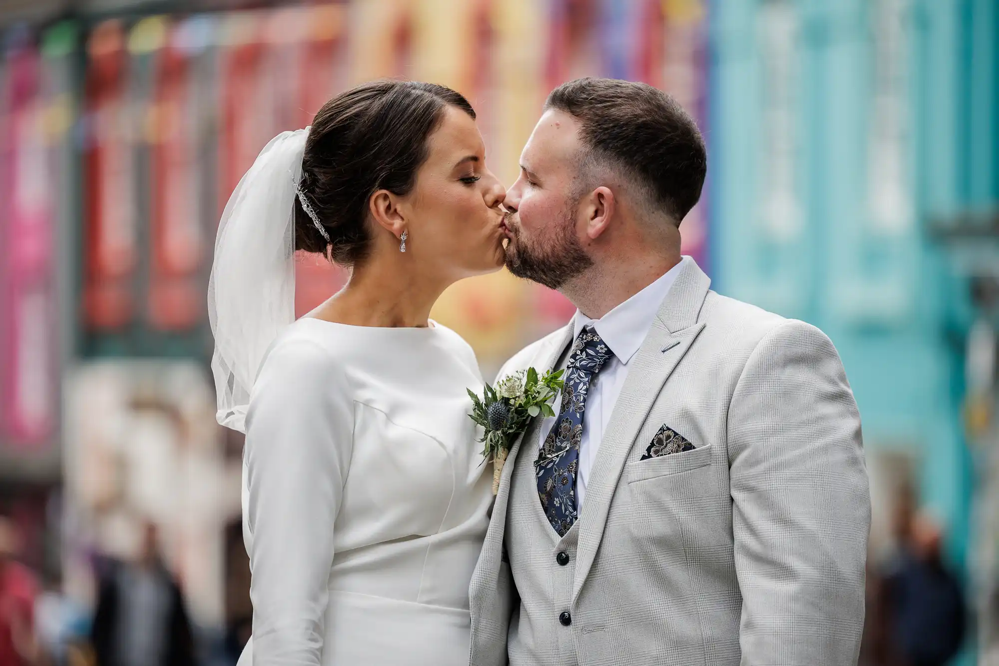 A bride in a white dress and veil kisses a groom in a light grey suit with a floral tie. Both face each other on a colourful, blurred street. The groom wears a buttonhole, and the bride has her hair in an updo.