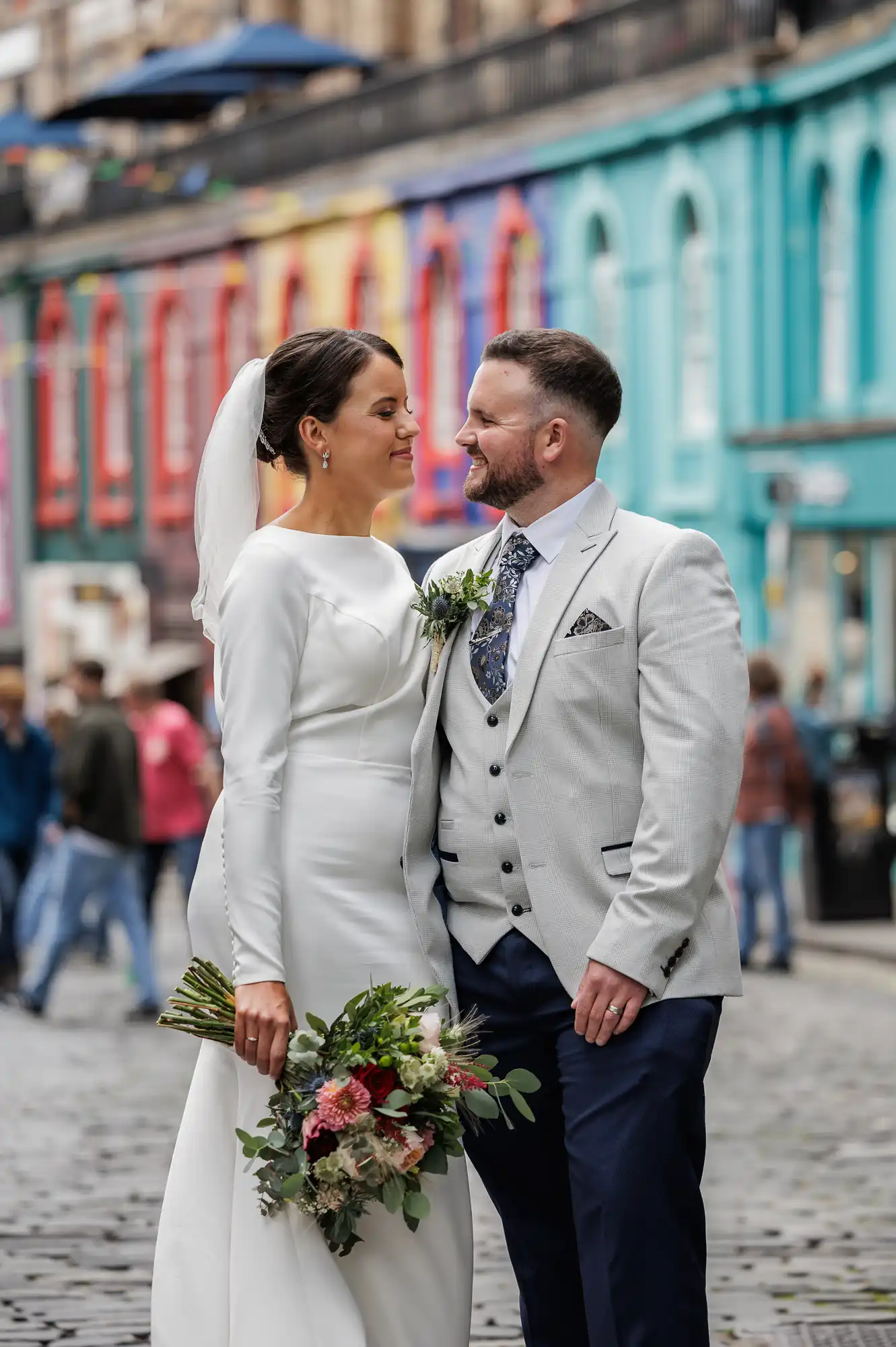 A bride in a long-sleeved white dress and veil stands smiling at her groom, who wears a light grey jacket, waistcoat, and patterned tie. She holds a bouquet of flowers. They stand on a cobbled street with colourful buildings in the background.