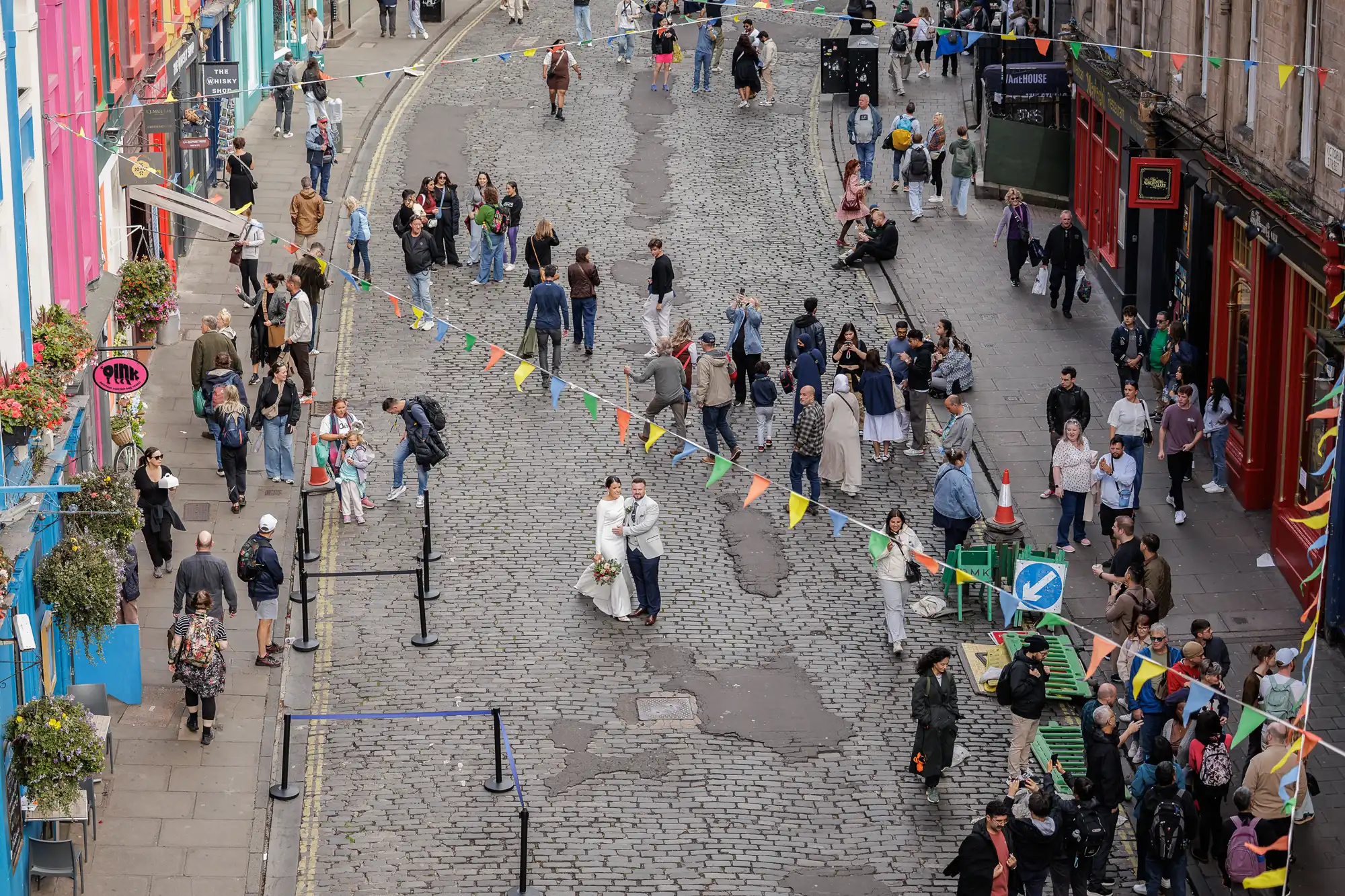 A bustling cobbled street scene features colourful buildings and festive bunting overhead. People walk, talk, and gather in groups, some carrying bags or taking photos, creating a lively, vibrant urban atmosphere.