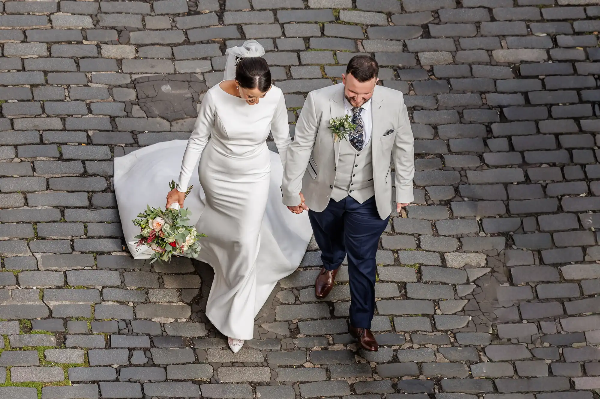 A bride in a long-sleeved white gown holding a bouquet walks hand-in-hand with a groom in a light grey jacket, white waistcoat, and navy trousers. They walk on cobblestone pavement, viewed from above, looking down and smiling.