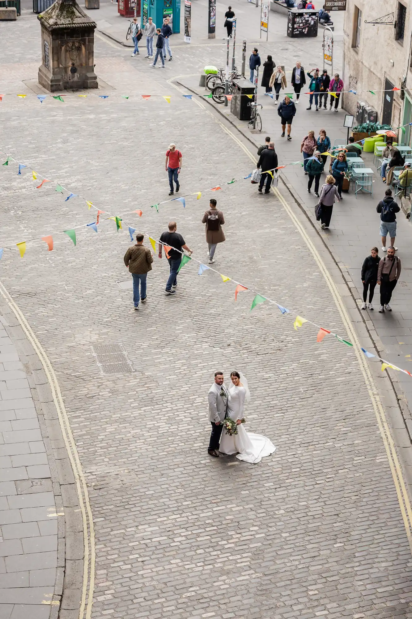 A bride and groom stand together on a cobbled street decorated with colourful bunting. People walk and gather on pavements around them, creating a lively urban scene. The couple is at the centre, smiling and holding hands, dressed in formal wedding attire.