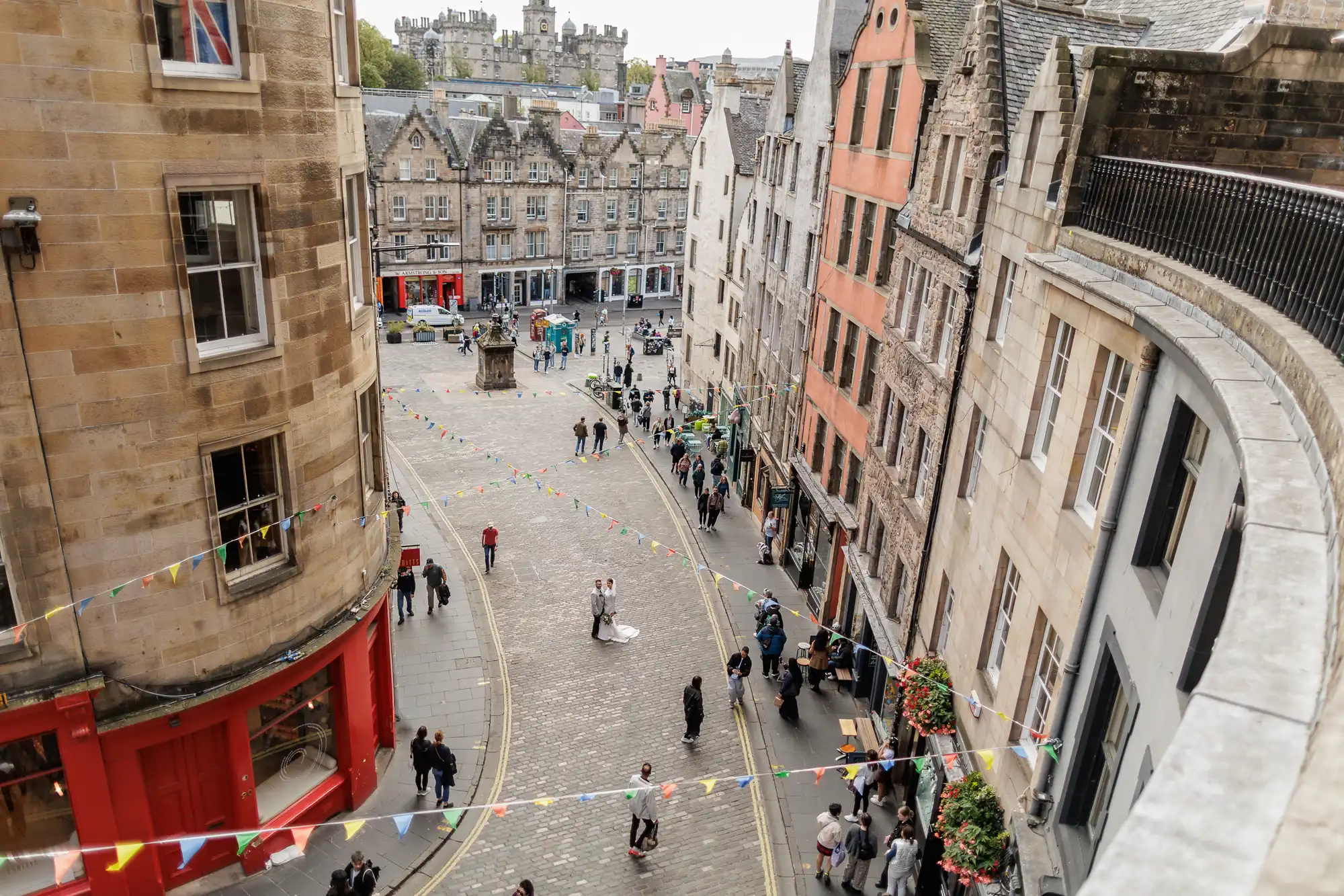 A cobblestone street curves between old stone buildings with shops and flats. Colourful bunting hangs above pedestrians. People stroll, some gather in small groups. The weather is cloudy. In the background, a castle-like structure sits on a hill.