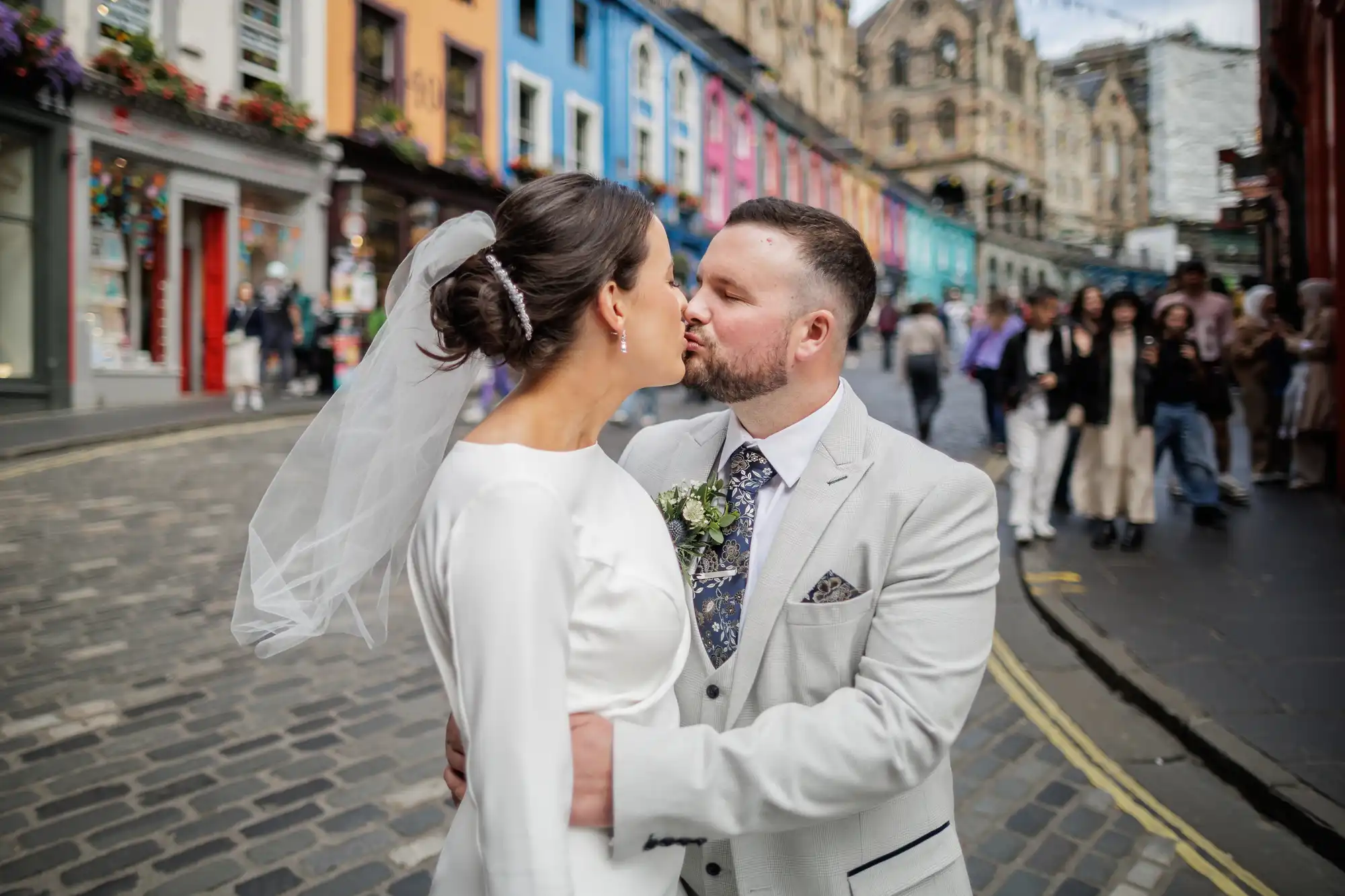 A bride in a white dress and veil faces a groom in a light suit as they embrace on a cobbled street lined with colourful buildings. The groom leans in to kiss her. People and historic architecture appear in the background.