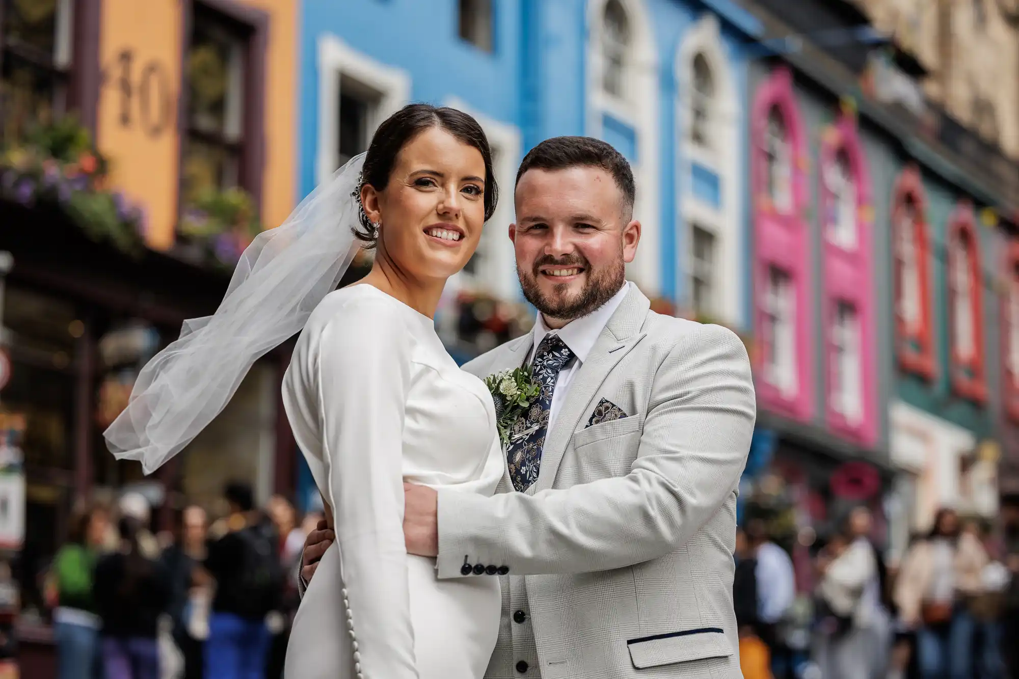 A smiling bride in a long-sleeved white gown and veil stands beside a groom in a light grey suit and floral tie. They embrace on a colourful street lined with blue, pink, and yellow buildings, with blurred people in the background.