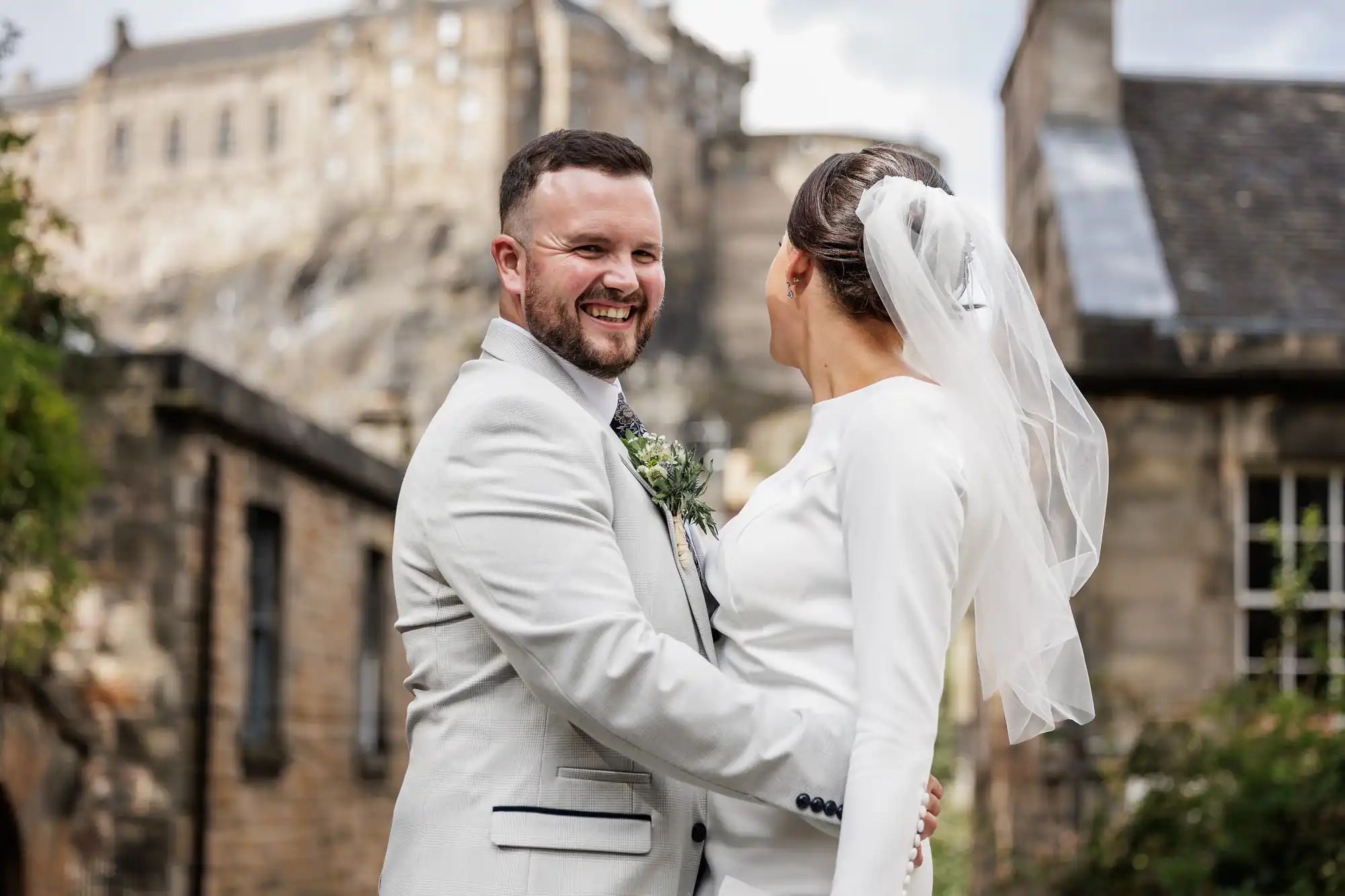 A groom in a light grey suit smiles joyfully whilst holding his bride, who wears a long-sleeved white dress and a veil. They stand outdoors beside old stone buildings with a large castle visible in the background.