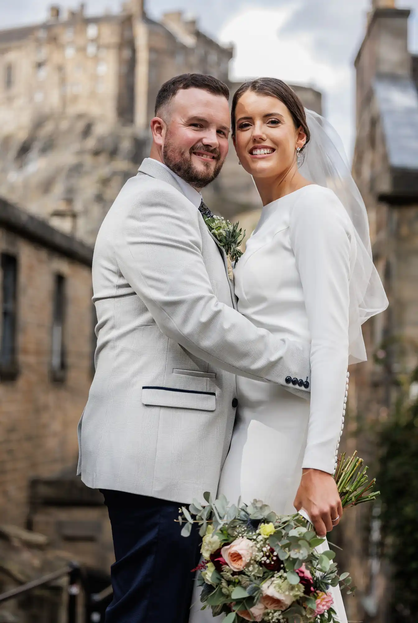 A bride in a white dress and veil stands beside a groom in a light grey suit. They smile and embrace outdoors, with a stone building and bouquet of flowers in the foreground, and a castle blurred in the background.