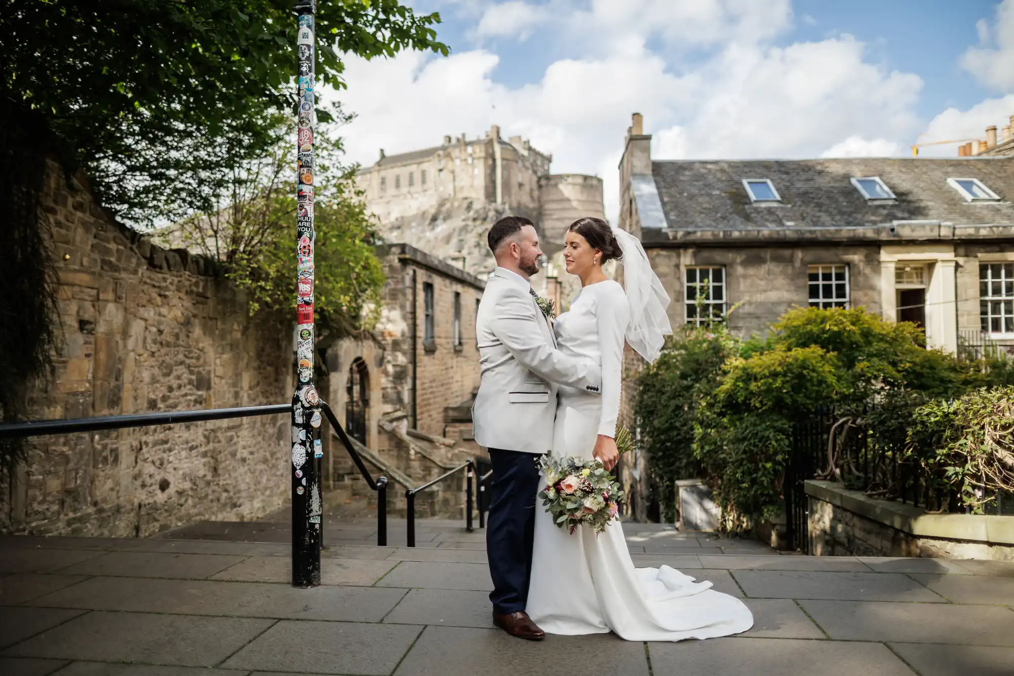 A bride and groom stand close together on a stone path outdoors, facing each other. The bride holds a bouquet and wears a white dress with a veil, while the groom wears a light jacket. Historic stone buildings and a castle appear in the background under a blue sky.