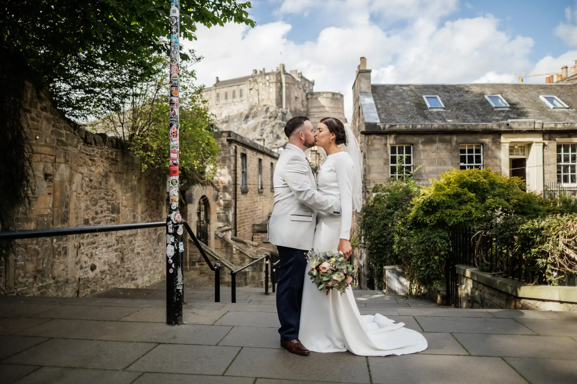 A bride and groom share a kiss outdoors on a stone walkway, with the bride holding a bouquet of flowers. They stand in front of old stone buildings and greenery, with a sticker-covered pole nearby and a large castle visible in the background under a blue sky.