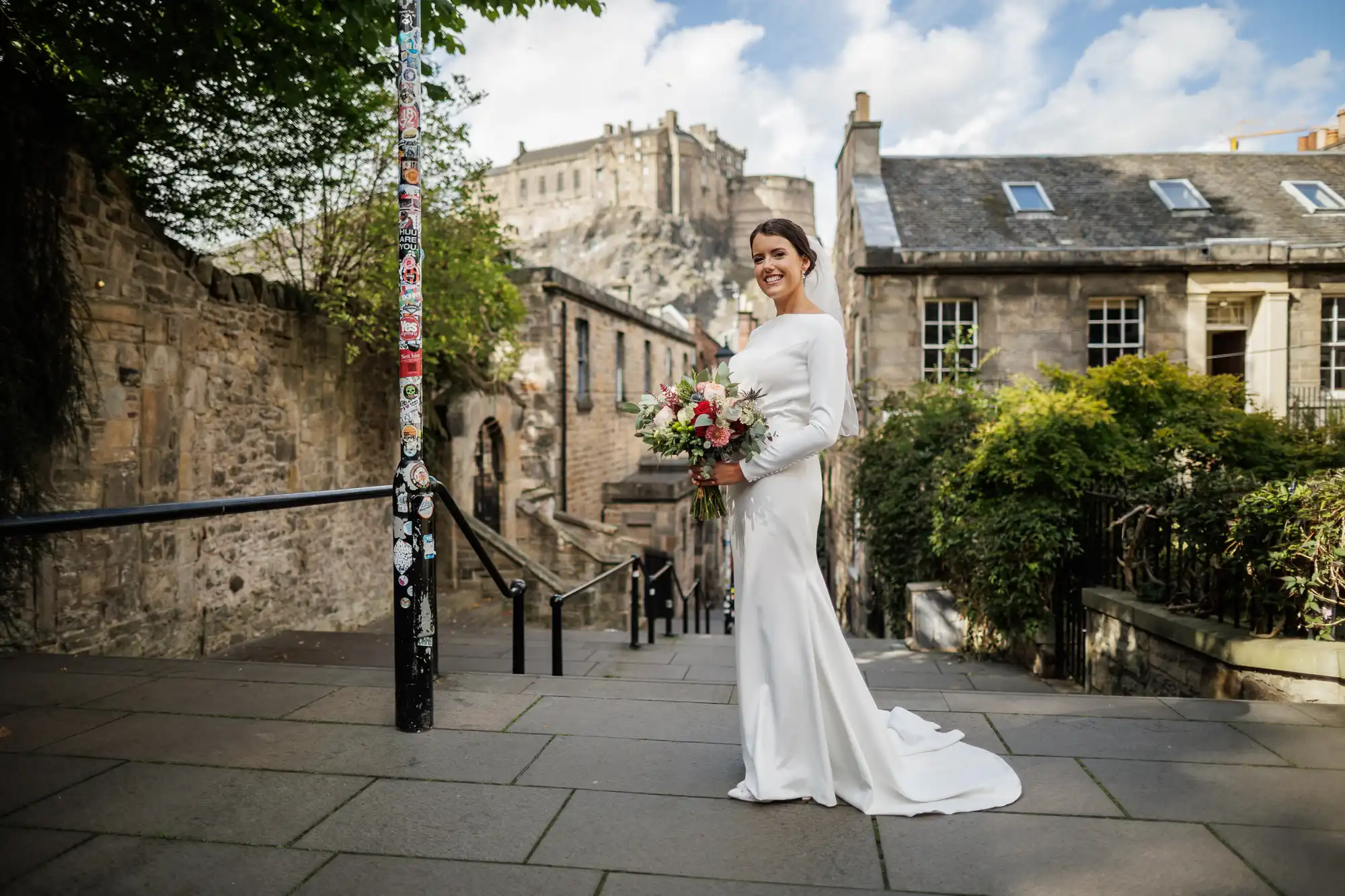 A smiling bride in a long-sleeved white gown holds a bouquet on a stone path, with historic buildings and Edinburgh Castle in the background under a partly cloudy sky. A pole covered in stickers stands to her left.