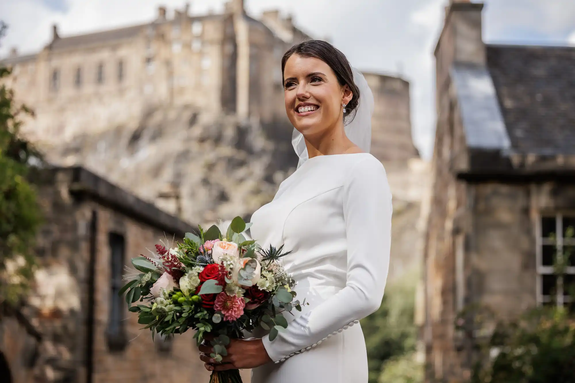 A smiling bride in a long-sleeved white dress holds a colourful bouquet of flowers. She stands outdoors between stone buildings, with a historic castle blurred in the background under a partly cloudy sky.