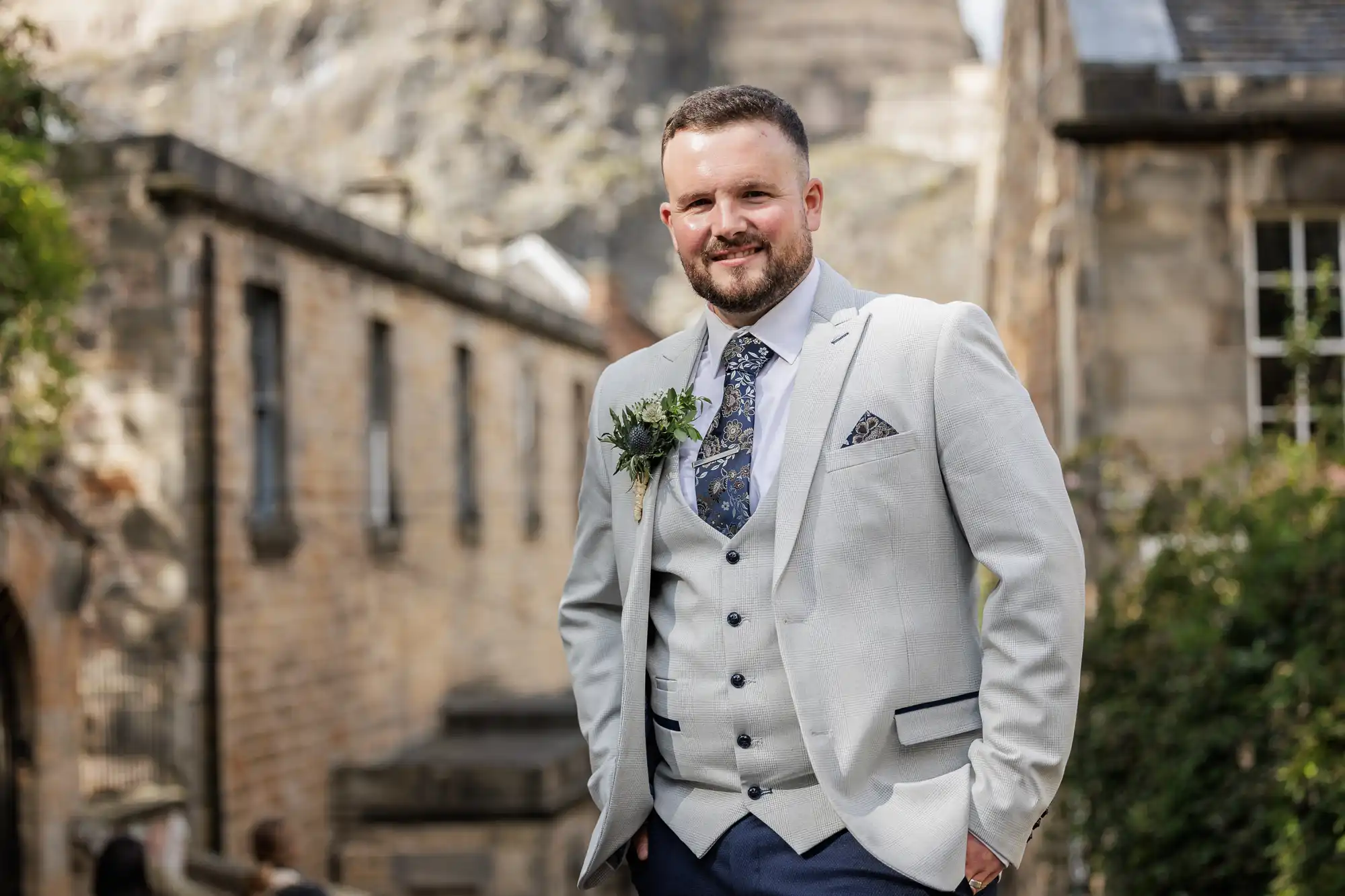 A smiling man with a beard stands outdoors, wearing a light grey suit, matching waistcoat, patterned tie, and pocket square. He has a buttonhole on his lapel. Behind him are old stone buildings and greenery, giving the setting a historic, rustic feel.