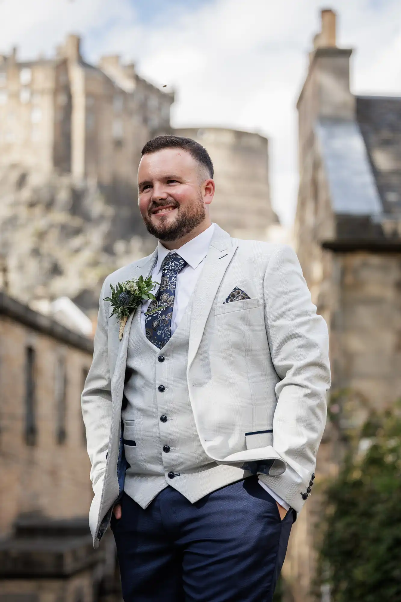 A smiling man with short brown hair and a beard wears a light grey suit jacket, matching waistcoat, white shirt, navy tie, and navy trousers. He has a floral buttonhole and stands outdoors among old stone buildings, with a castle visible in the background.