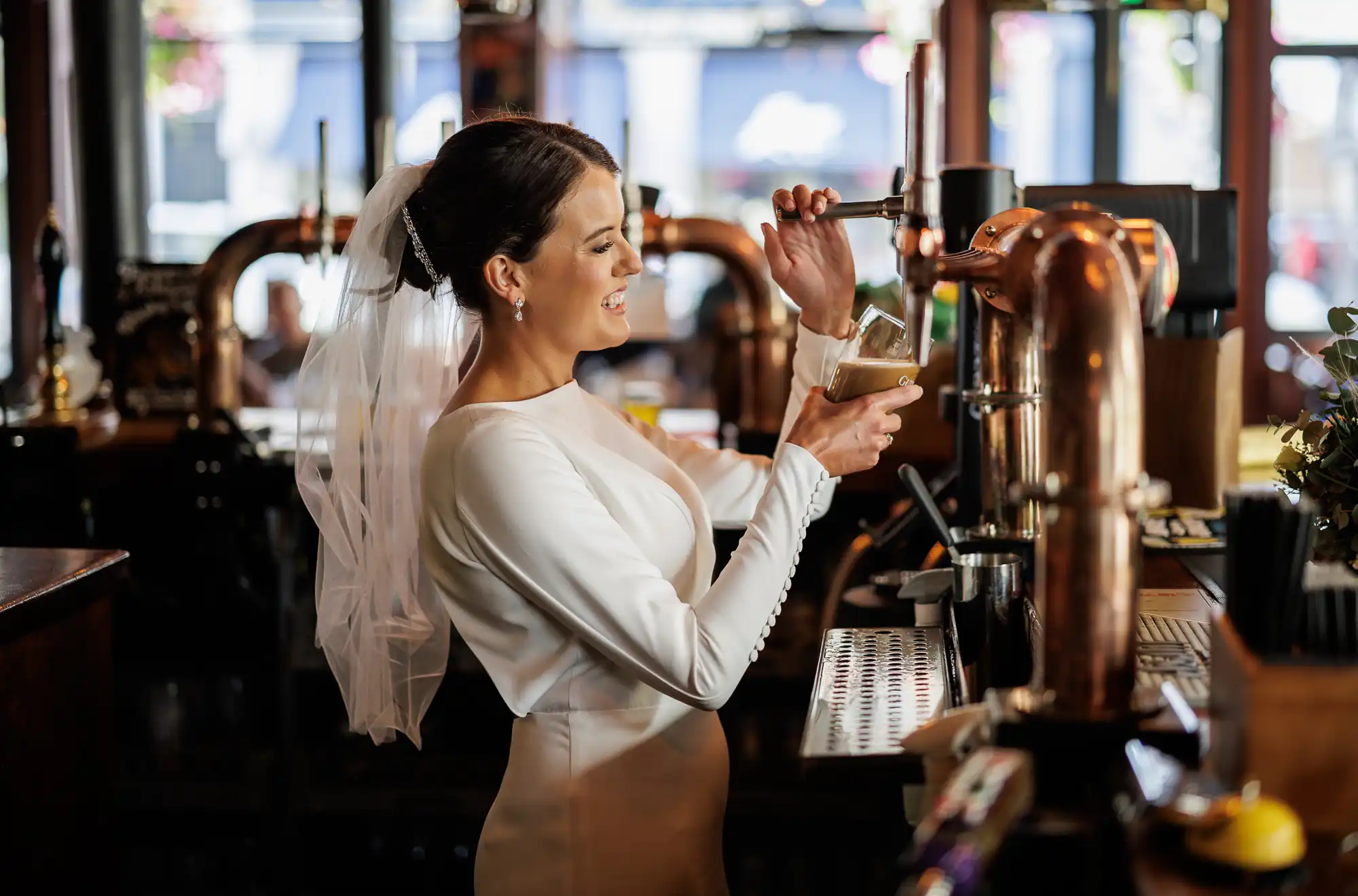 A bride in a white dress and veil smiles as she pours a drink from a beer tap behind a bar, holding a glass. The bar has copper taps and a modern interior, with blurred windows and floral decorations in the background.