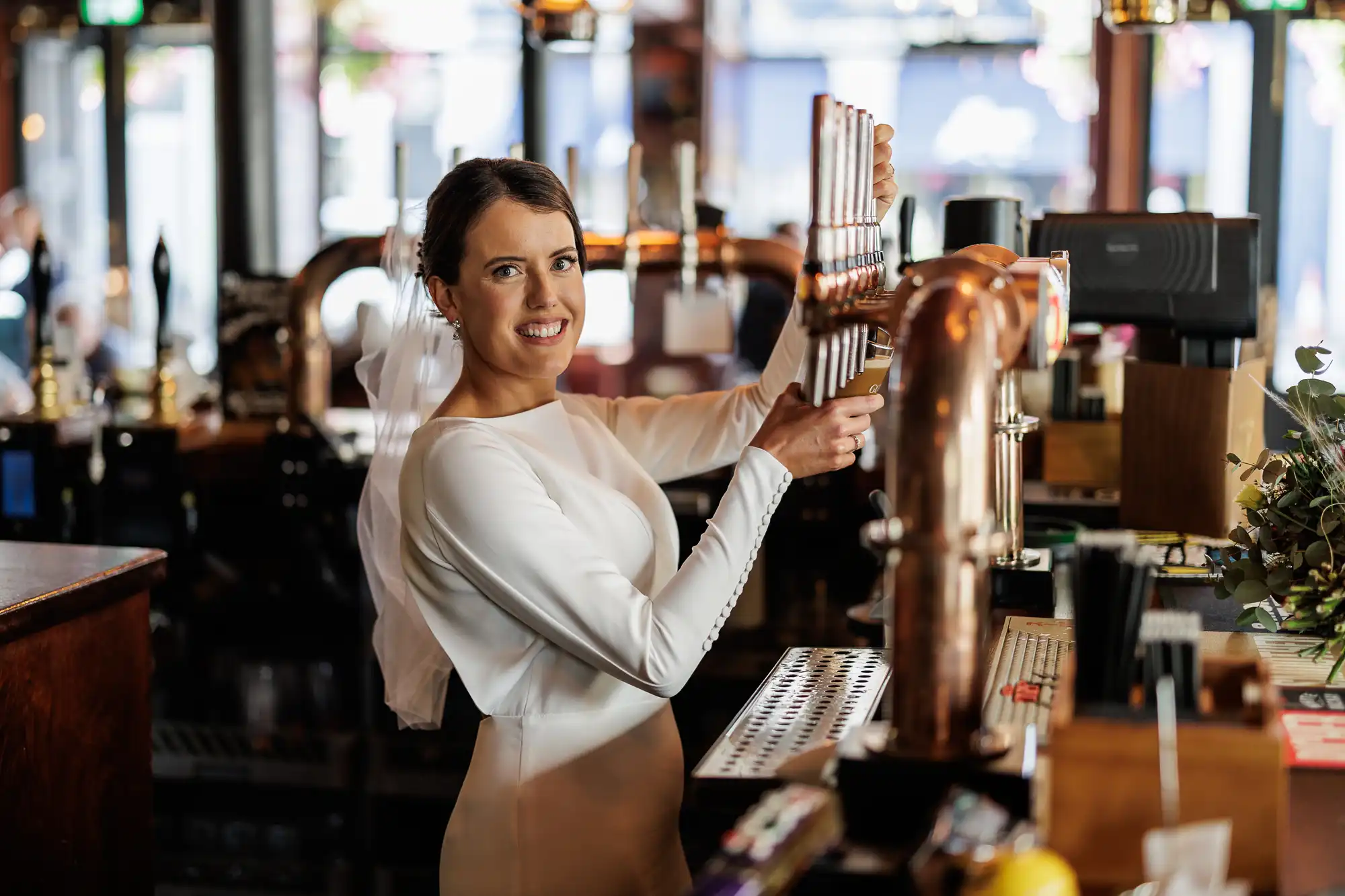 A bride in a long-sleeved white wedding dress and veil stands behind a bar, smiling at the camera while pulling a beer pump. The bar has copper pipes, glasses, and plants, with a bright and lively interior in the background.