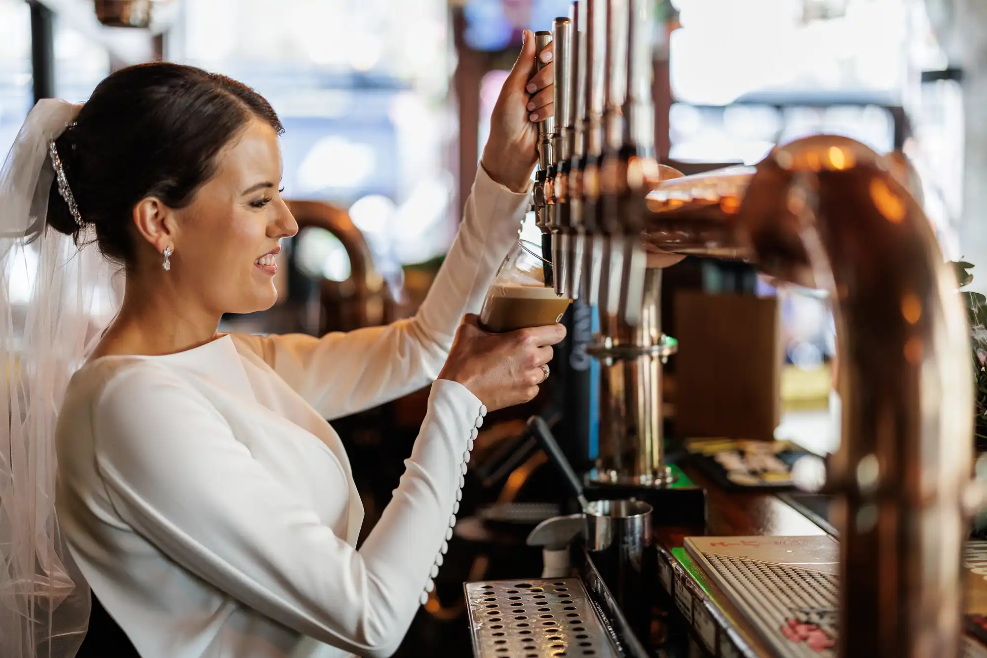 A smiling bride in a white dress and veil pours a pint of stout from a tap behind a bar, surrounded by copper pipes and bar equipment, with soft daylight shining in from the window.