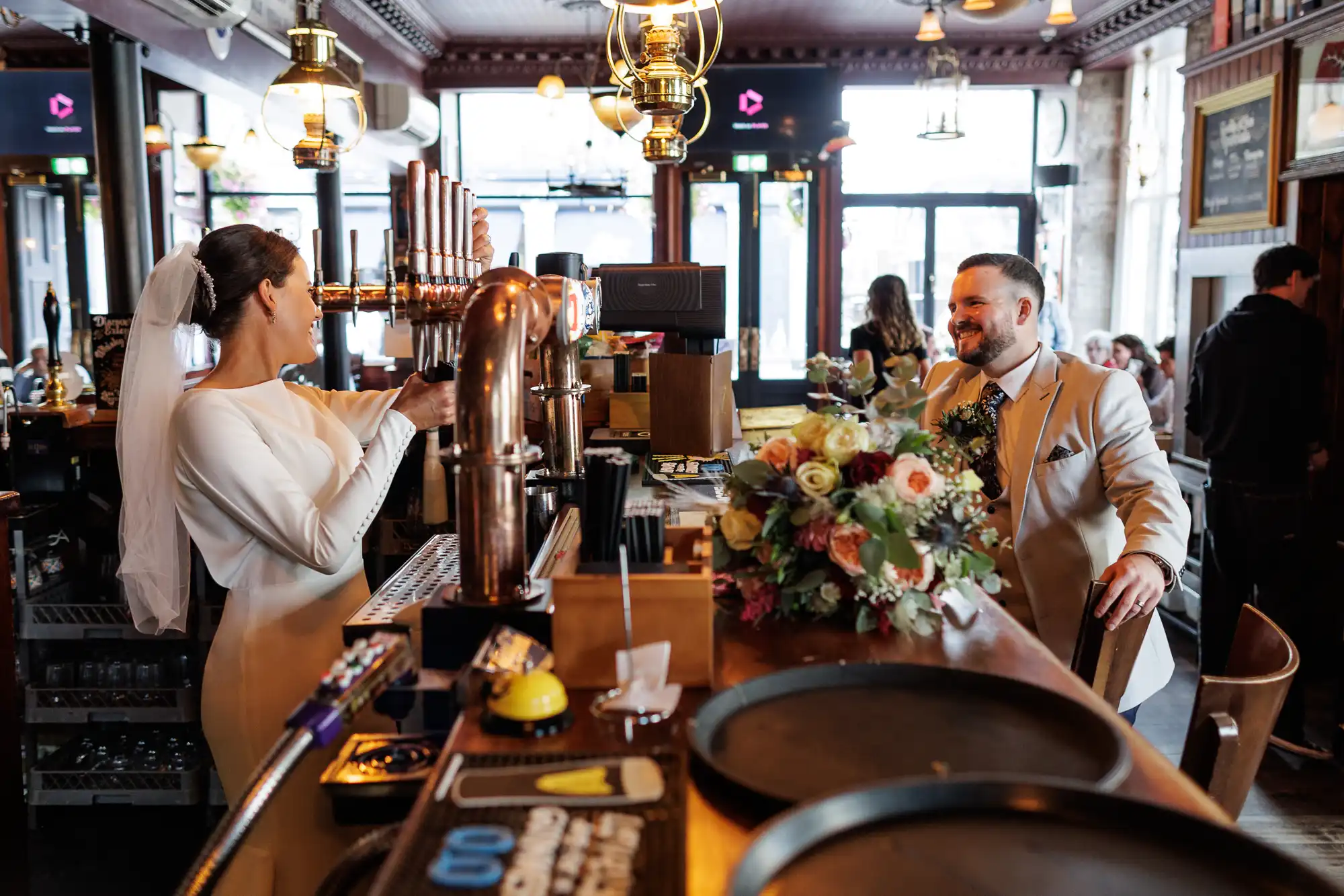 A bride in a white dress and veil pours a beer from a tap behind a bar, smiling at a groom in a beige suit sitting at the bar. A bouquet sits on the bar; the scene is warmly lit with modern fittings and patrons in the background.