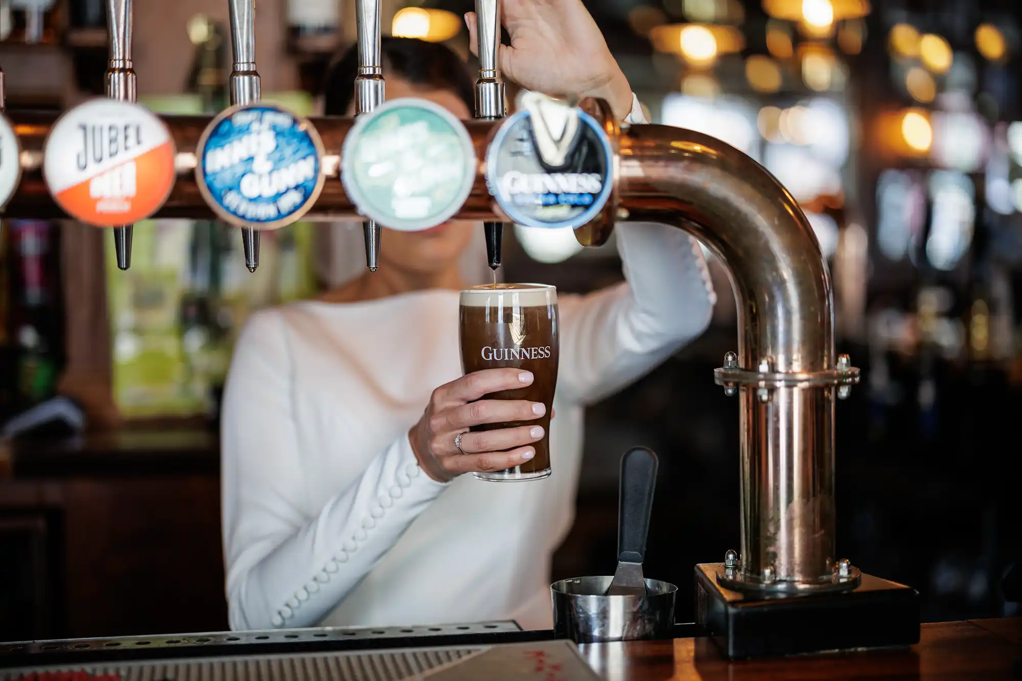 A person in a white long-sleeve shirt pours a pint of Guinness from a tap at a bar, holding the glass under the spout. The background shows blurred bar lights and other beer taps with colourful labels.