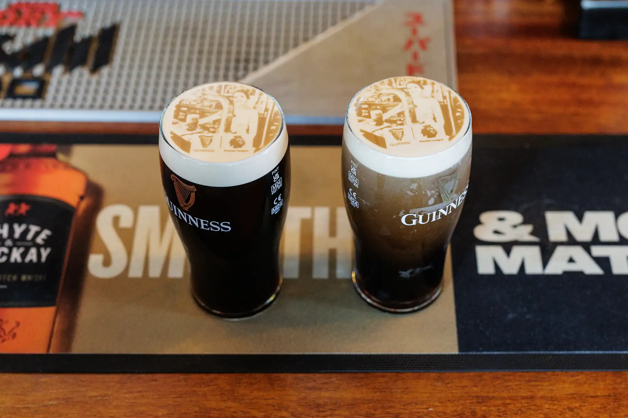 Two pint glasses of Guinness beer sit on a bar mat. Each has a creamy white foam head with an intricate printed image on top. The bar surface is wooden, with parts of branded mats and drink adverts visible in the background.