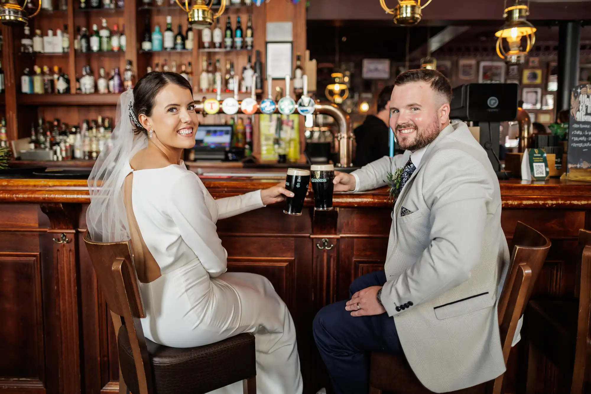 A bride in a white dress and veil and a groom in a light suit sit at a wooden bar, smiling at the camera and holding pints of dark beer, surrounded by bottles and taps in a warmly lit pub setting.