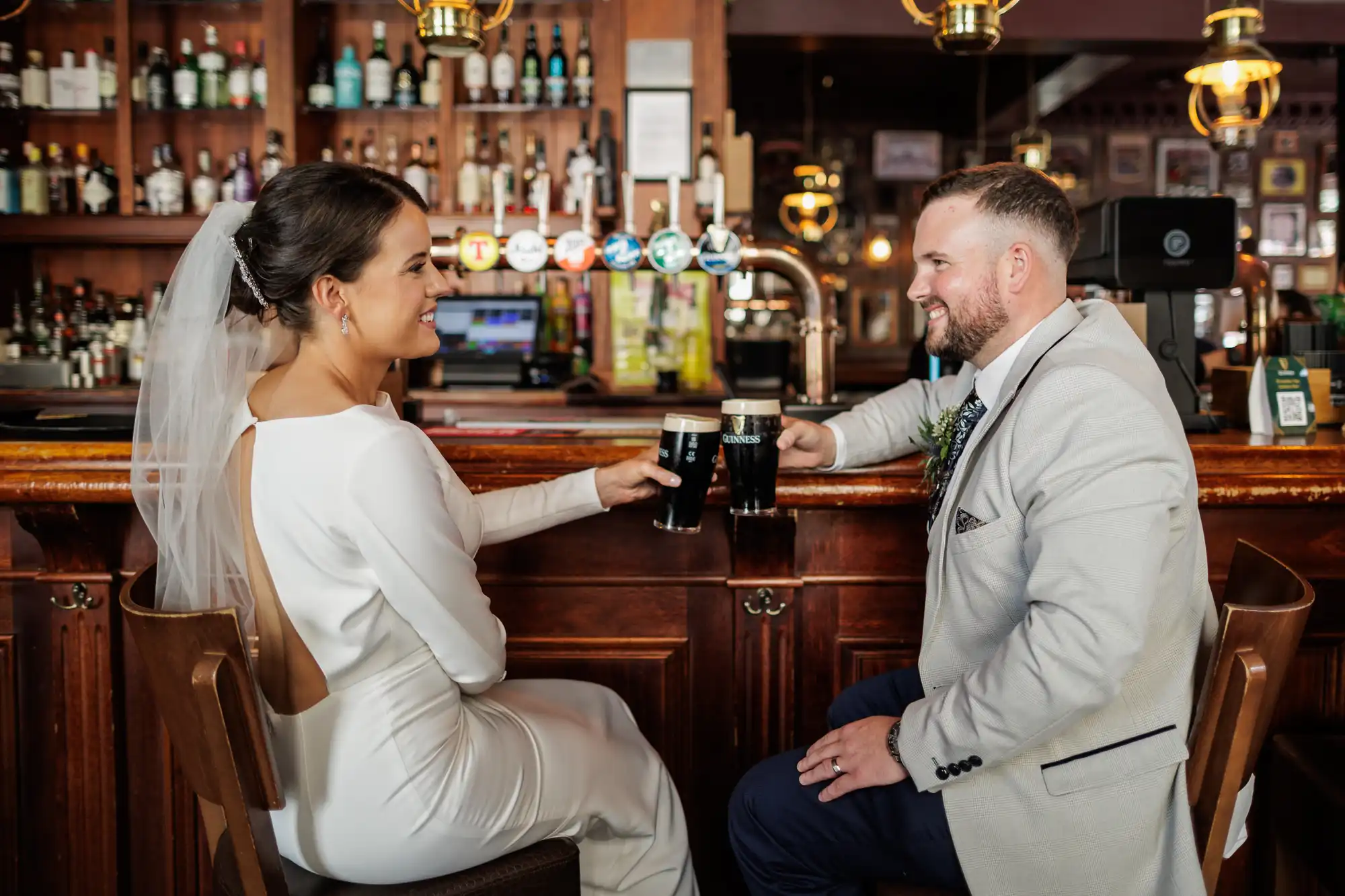 A bride in a white dress and veil and a groom in a light suit sit at a wooden bar, smiling at each other and toasting with pints of dark beer. Bottles and pumps line the bar shelves in the background, creating a warm, celebratory atmosphere.