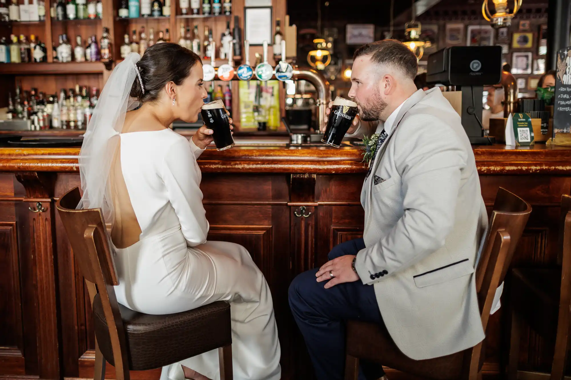 A bride in a white dress and veil and a groom in a light suit sit at a wooden bar, facing each other, each holding a pint of dark beer. The bar’s shelves behind them are stocked with bottles and glasses, creating a cosy atmosphere.