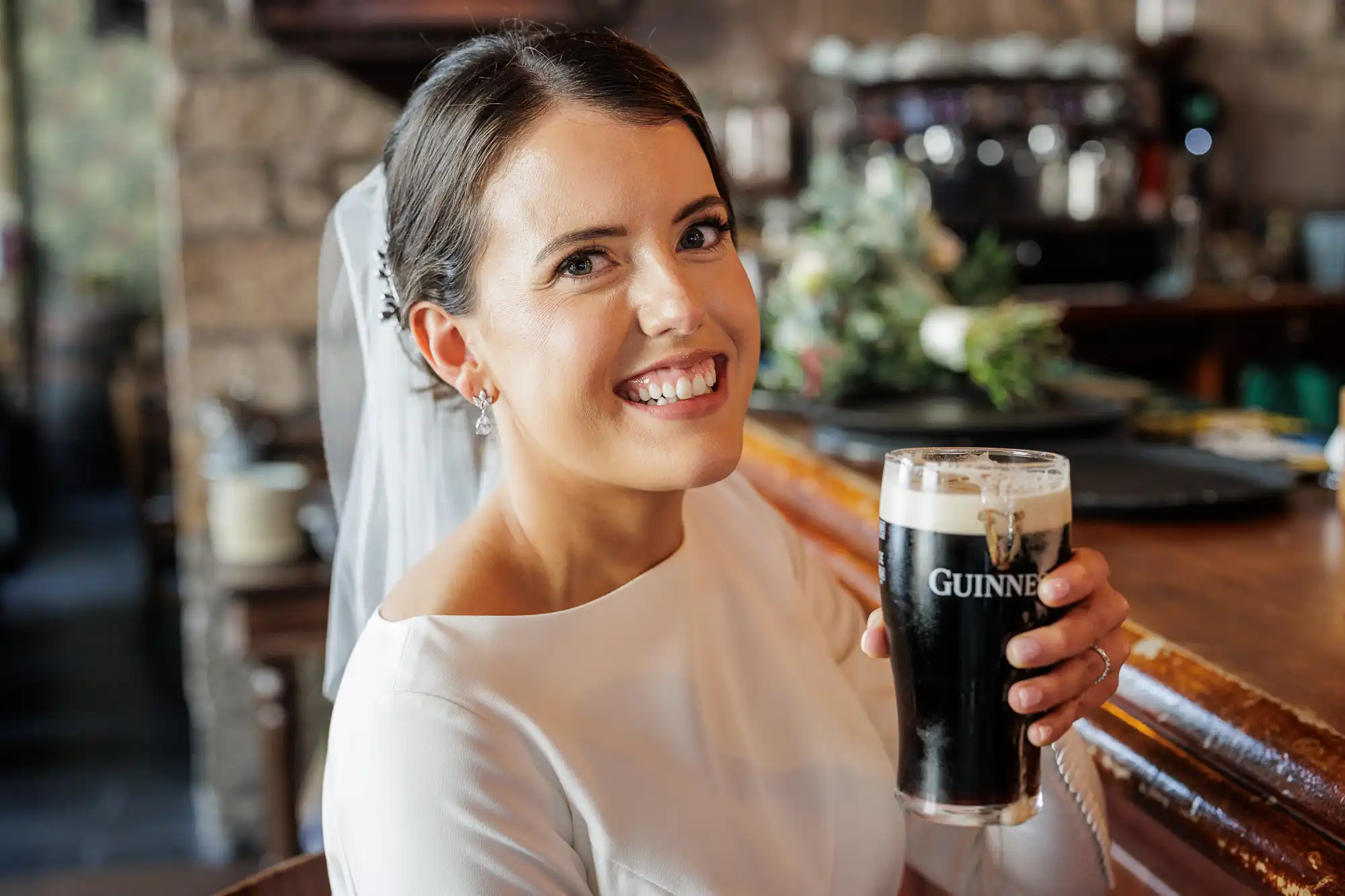 A smiling bride in a white dress and veil sits at a wooden bar, holding a pint of Guinness. She looks at the camera with a cheerful expression. The background has blurred bar details and a bouquet of flowers on the counter.
