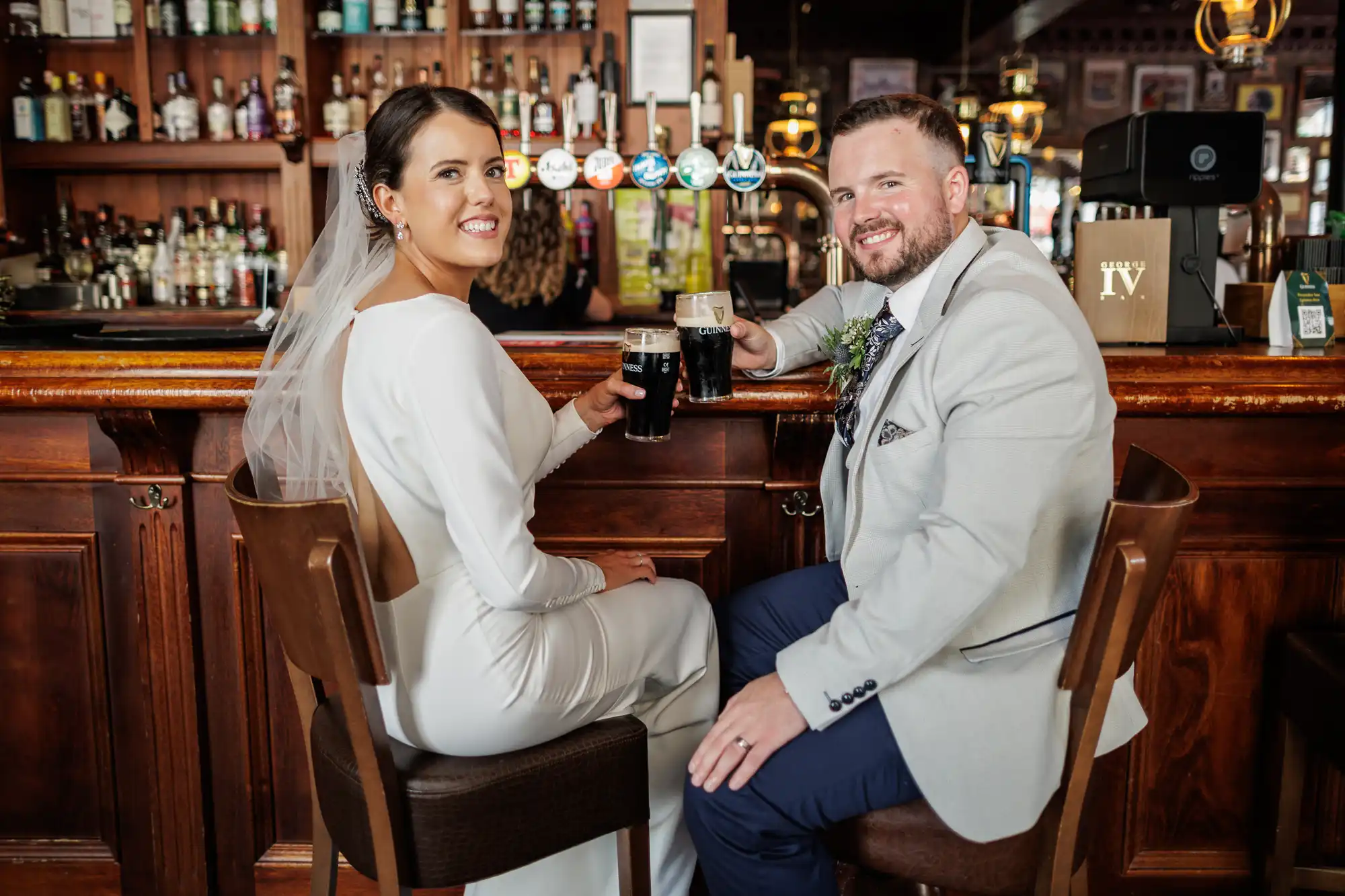 A bride in a white dress and veil and a groom in a light suit sit on bar stools at a pub, smiling at the camera and clinking pints of dark beer, with shelves of bottles and beer taps visible behind them.