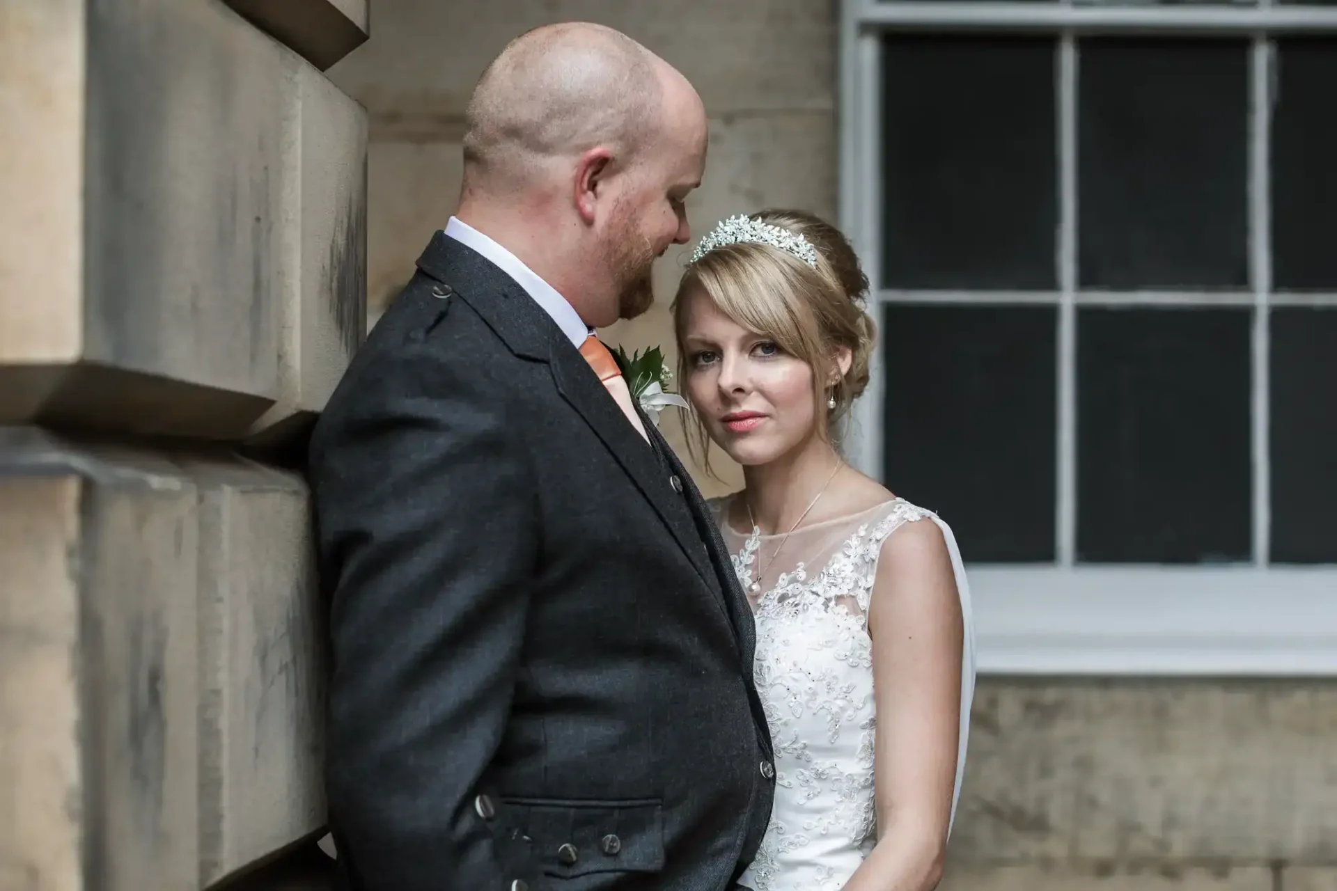 A bride and groom stand close together against a stone wall, with the groom looking at the bride and the bride facing the camera.