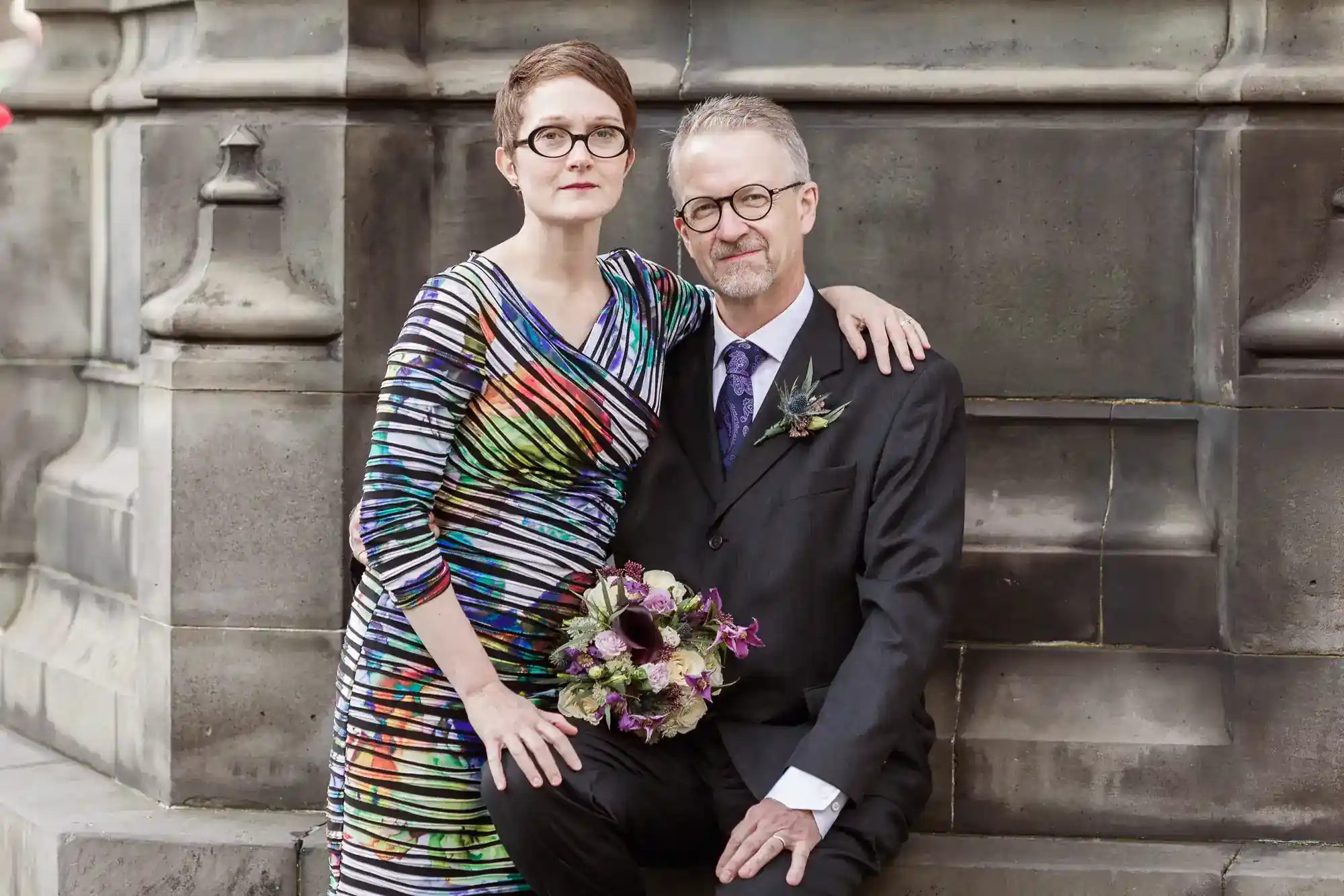 A woman in a colorful striped dress stands beside a man in a suit and tie; she holds a bouquet, and they pose together in front of a stone wall.