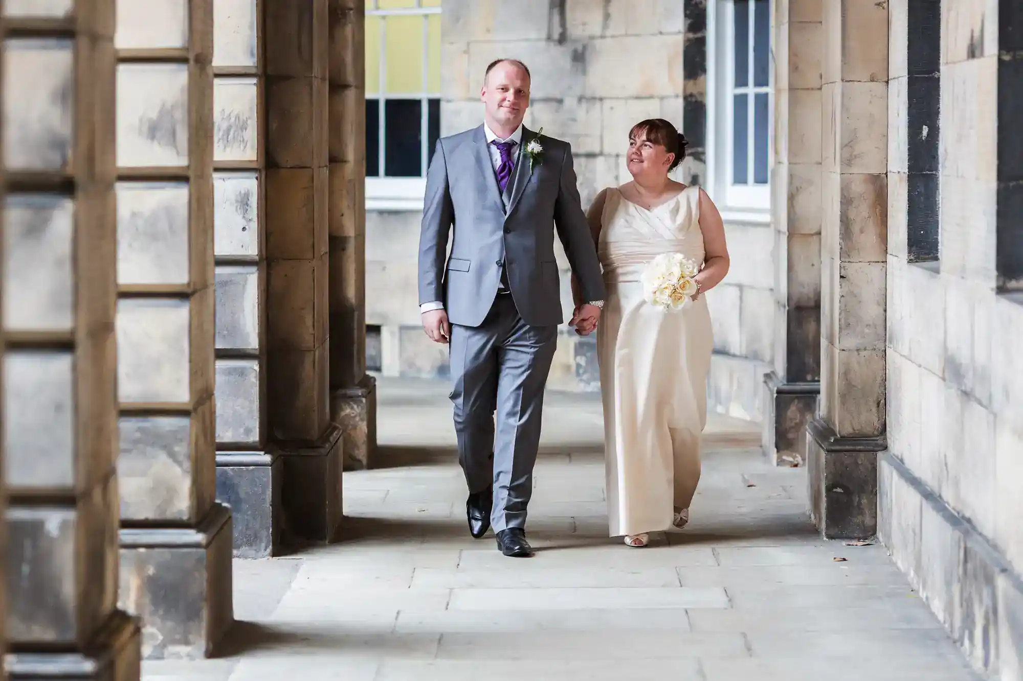 A couple dressed in formal attire walks hand in hand through a stone corridor, with the woman holding a small bouquet.