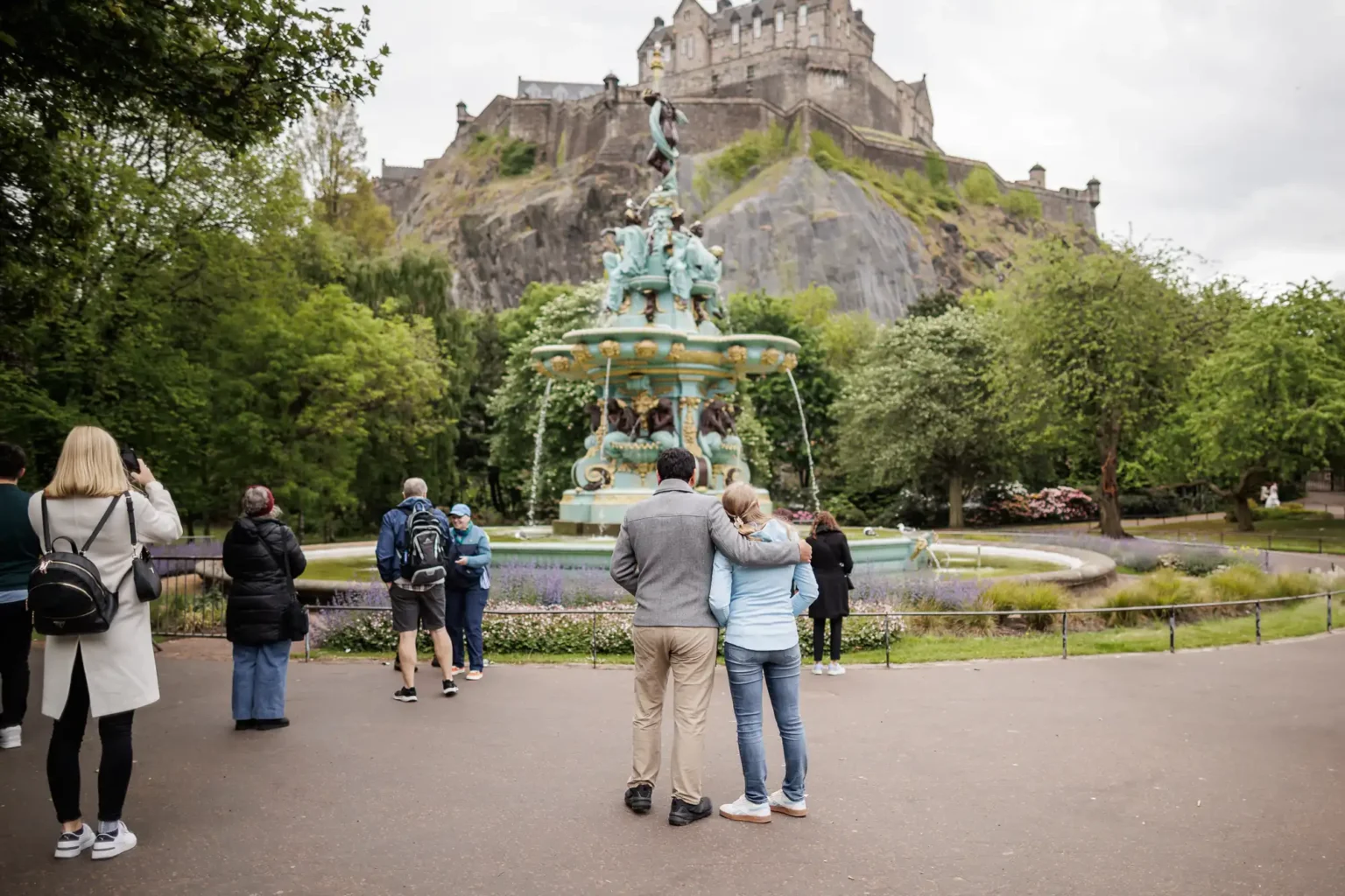 People stand around a decorative fountain in a park, with a castle on a hill in the background. Some are taking photos, while a couple stands close together in the foreground.
