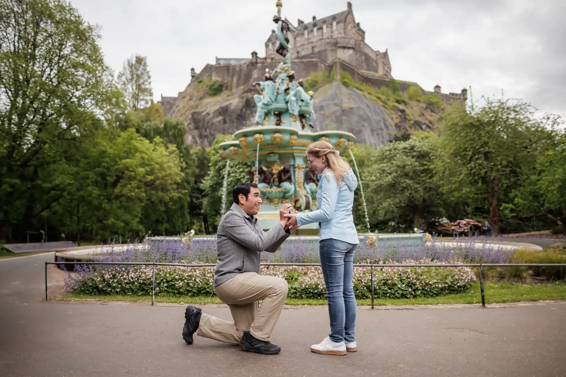 A man kneels and proposes to a woman in front of the Ross Fountain, with Edinburgh Castle visible in the background.