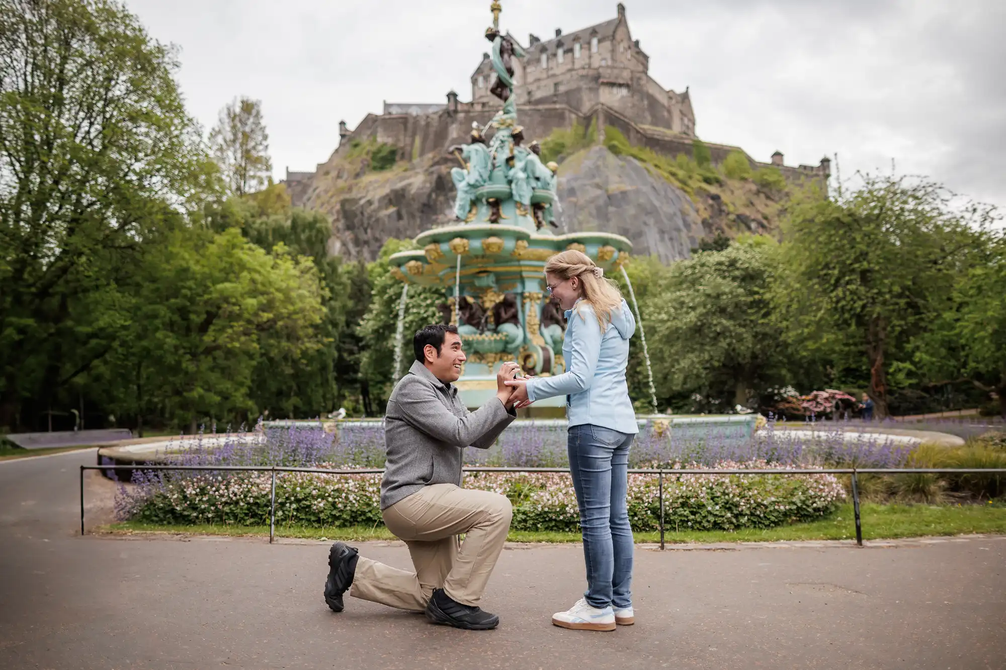 A man kneels and proposes to a woman in front of the Ross Fountain, with Edinburgh Castle visible in the background.