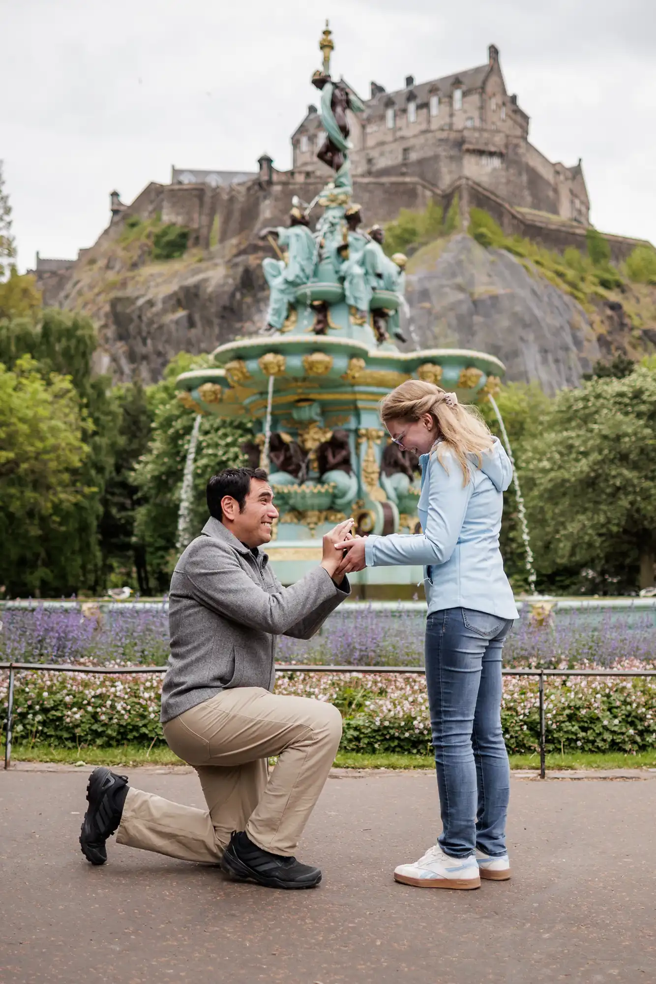 A man kneels and proposes to a woman in front of a decorative fountain with a historic castle on a hill in the background.