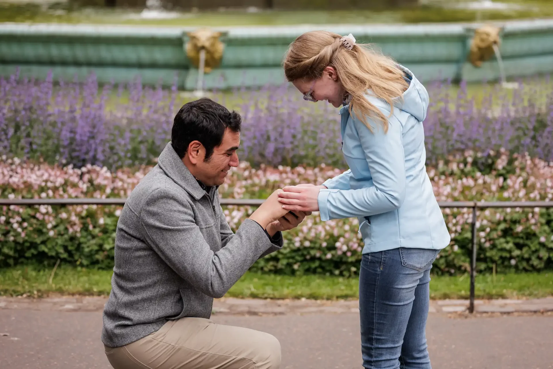 A man kneels and holds a ring as he proposes to a woman outdoors near a garden with flowers and a fountain in the background.