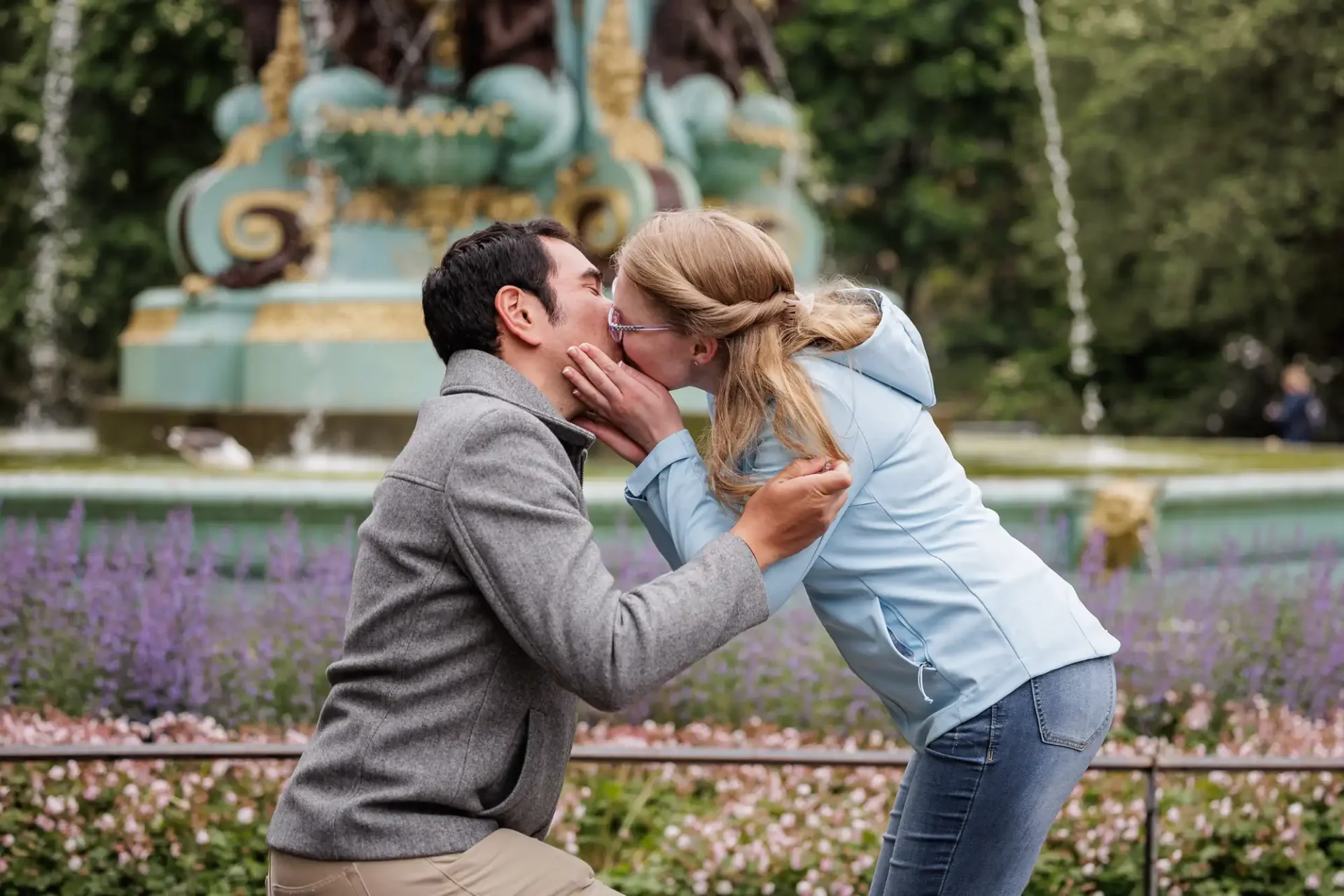 A man kneels and proposes to a woman in front of a decorative fountain; she leans in to kiss him, smiling, surrounded by flowers and greenery.