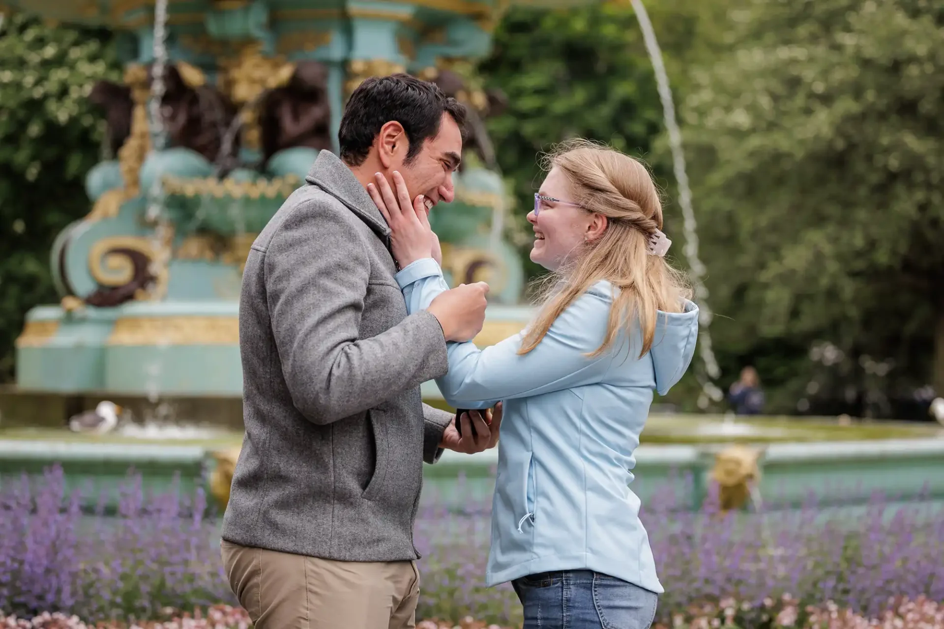 A man and woman stand facing each other in a park, smiling and touching faces, with a decorative fountain and flowers in the background.