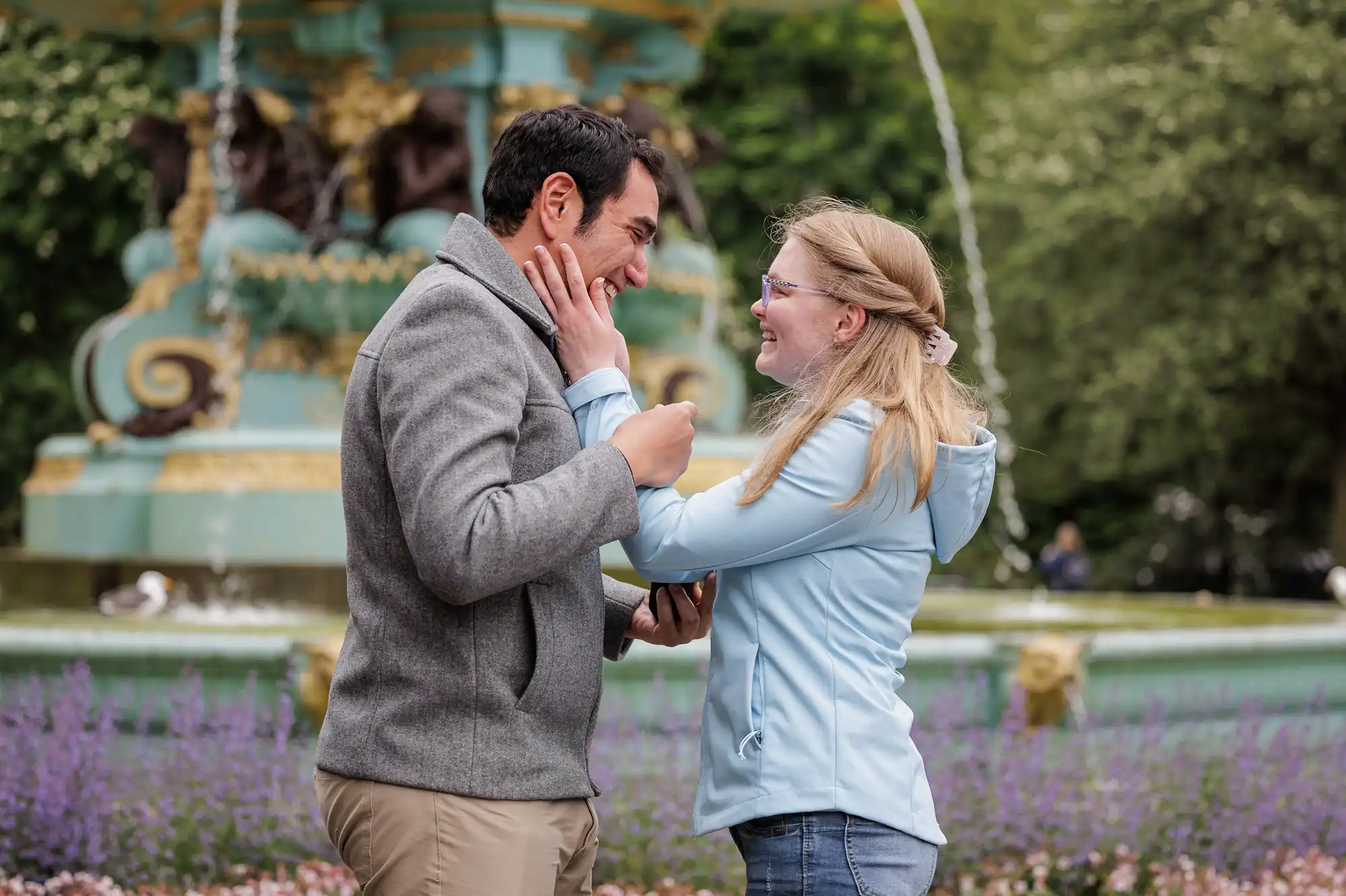 A man and woman stand facing each other in a park, smiling and touching faces, with a decorative fountain and flowers in the background.