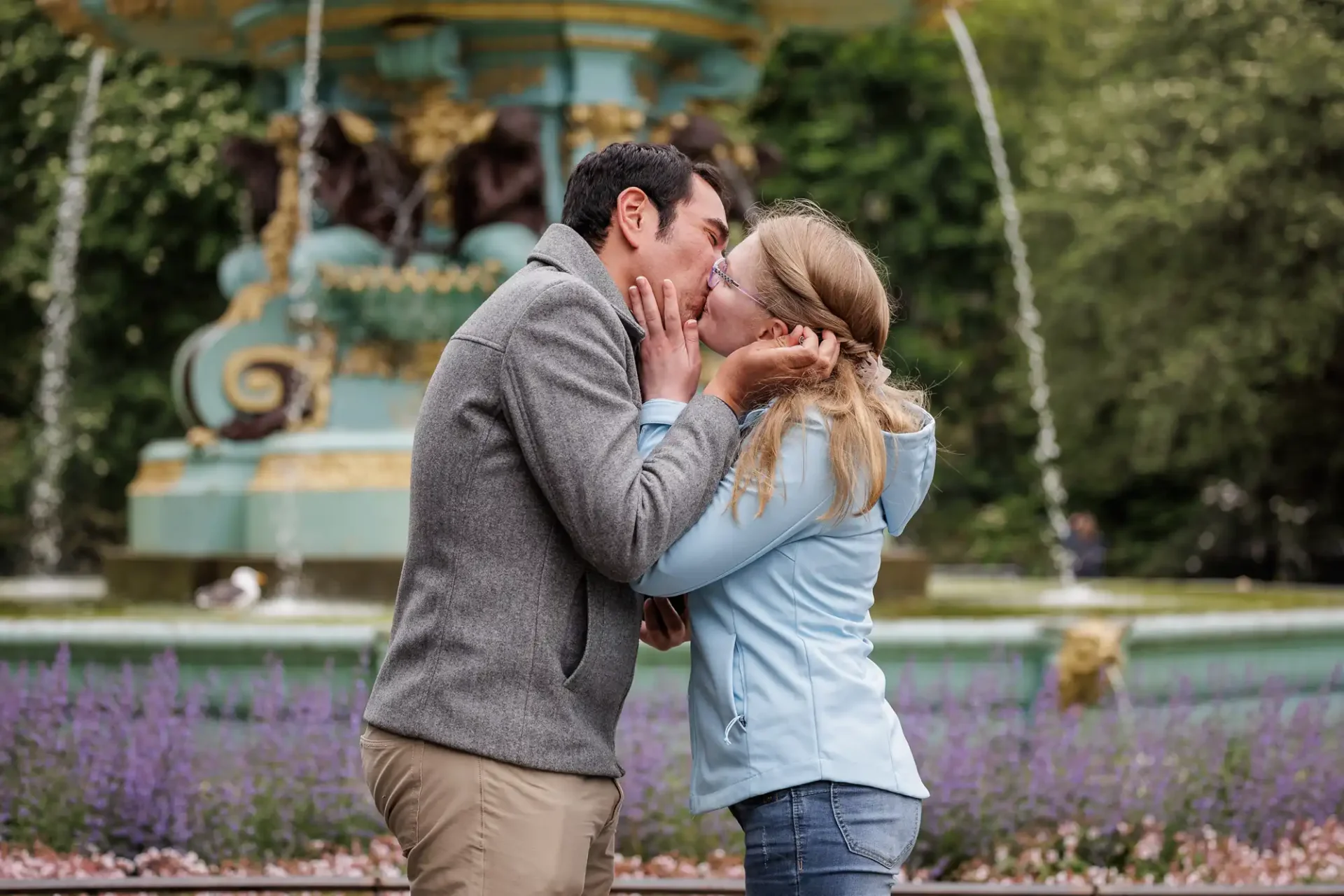 A man and woman kiss in front of a decorative fountain in a park, surrounded by greenery and purple flowers.