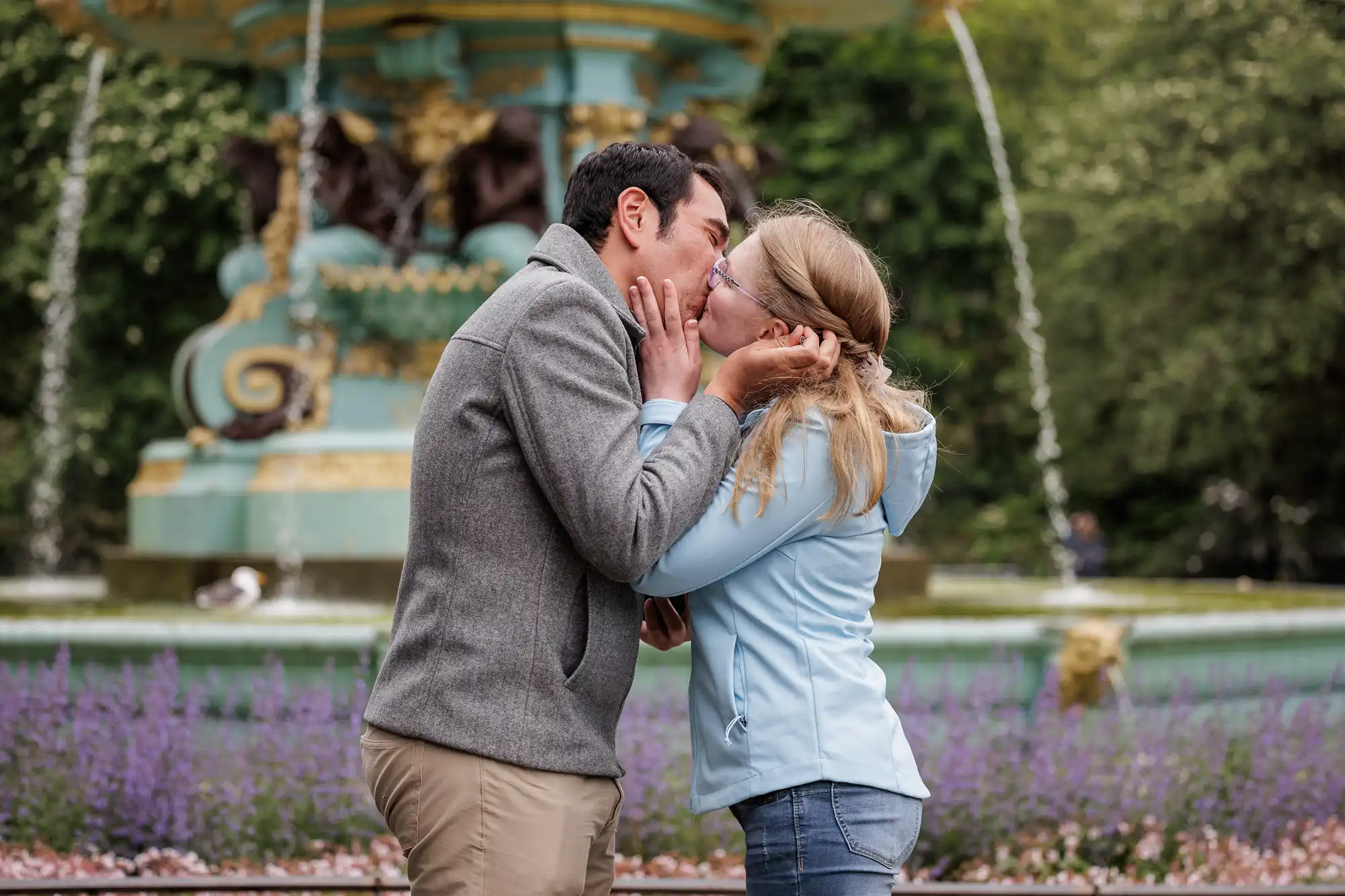 A man and woman kiss in front of a decorative fountain in a park, surrounded by greenery and purple flowers.