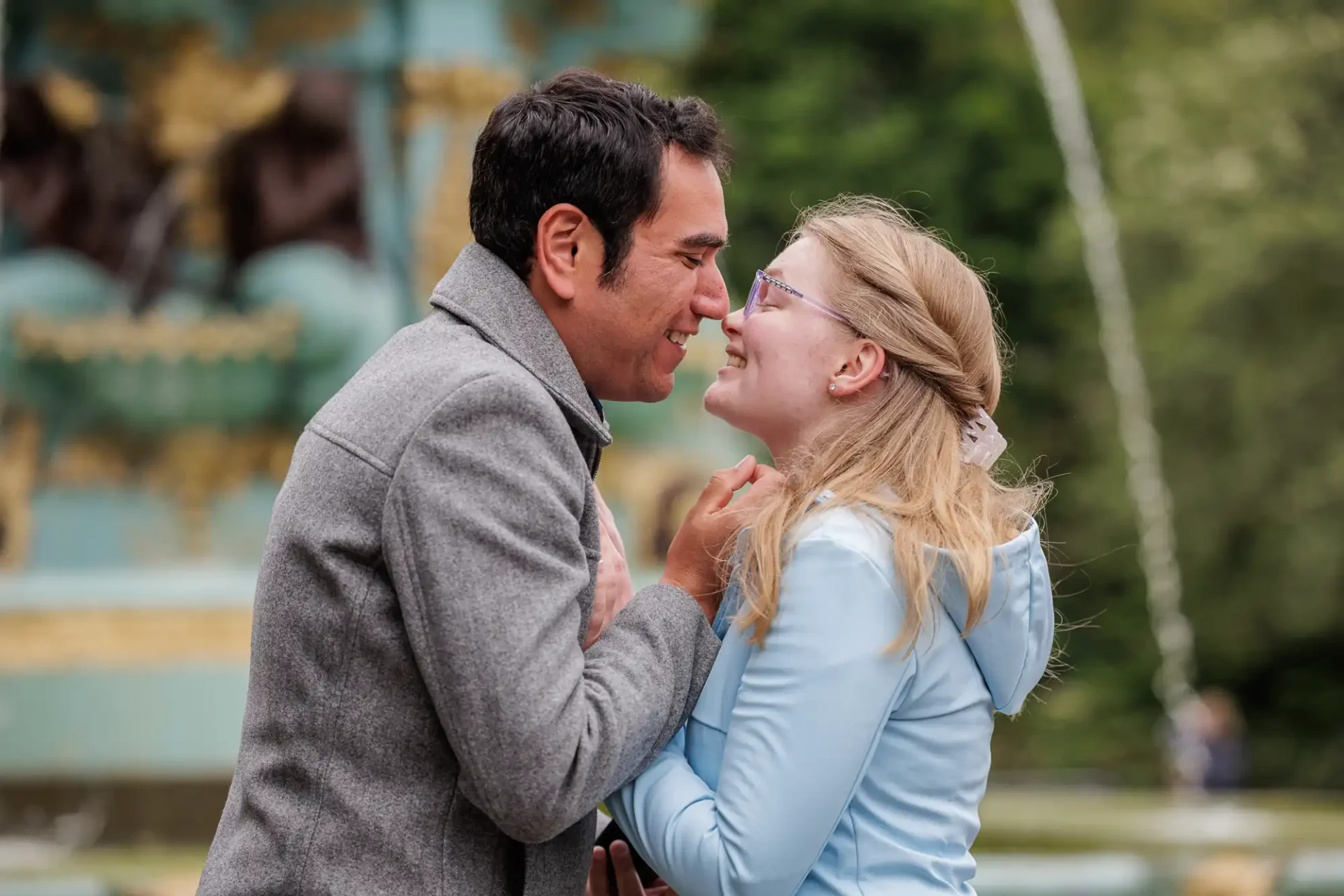 A man and a woman stand close together outdoors, smiling at each other and holding hands near a decorative fountain.
