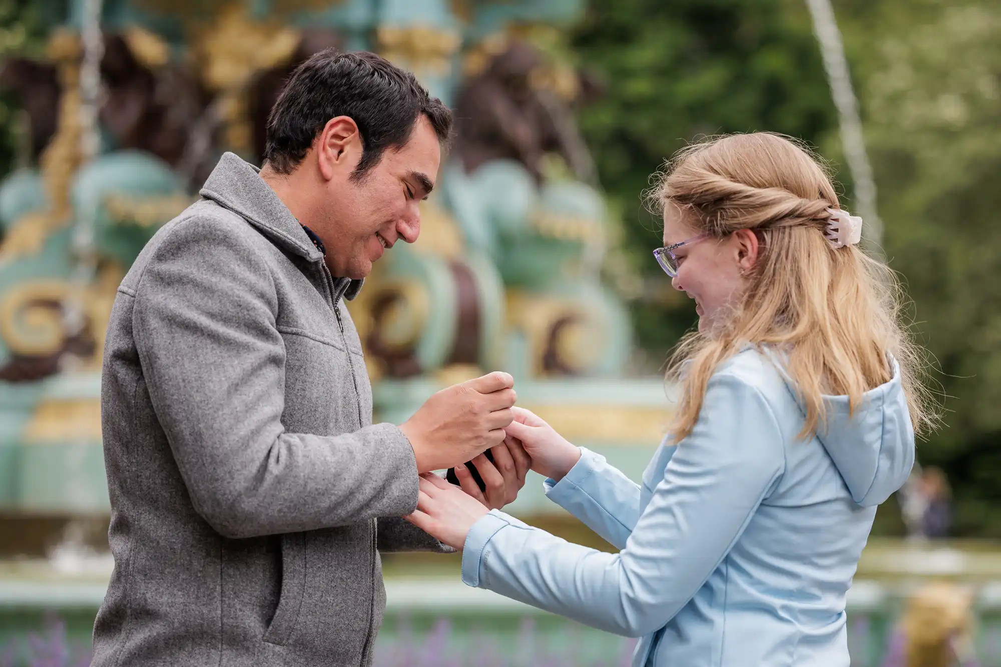 A man places a ring on a woman’s finger outdoors, both smiling, with a decorative fountain and greenery in the background.