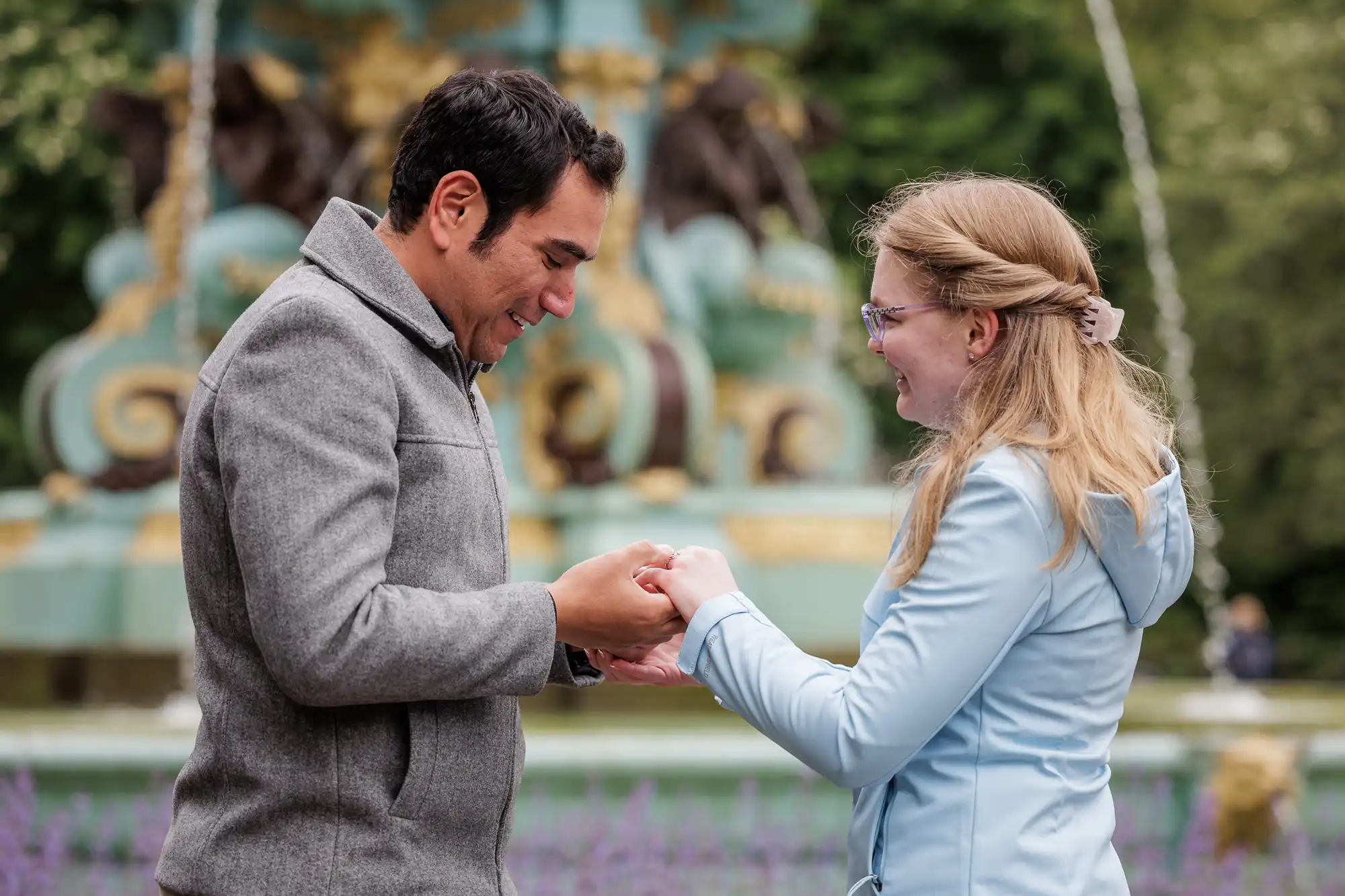 A man and woman stand facing each other outdoors, holding hands and smiling, with a decorative fountain in the background.