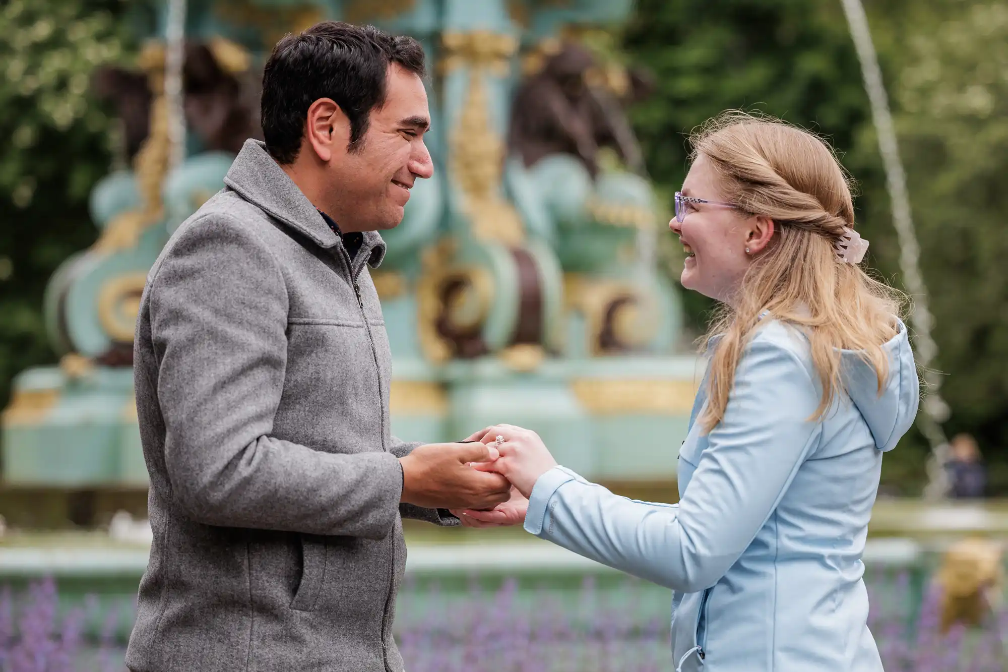 A man and woman stand facing each other outdoors, holding hands and smiling near a decorative fountain.