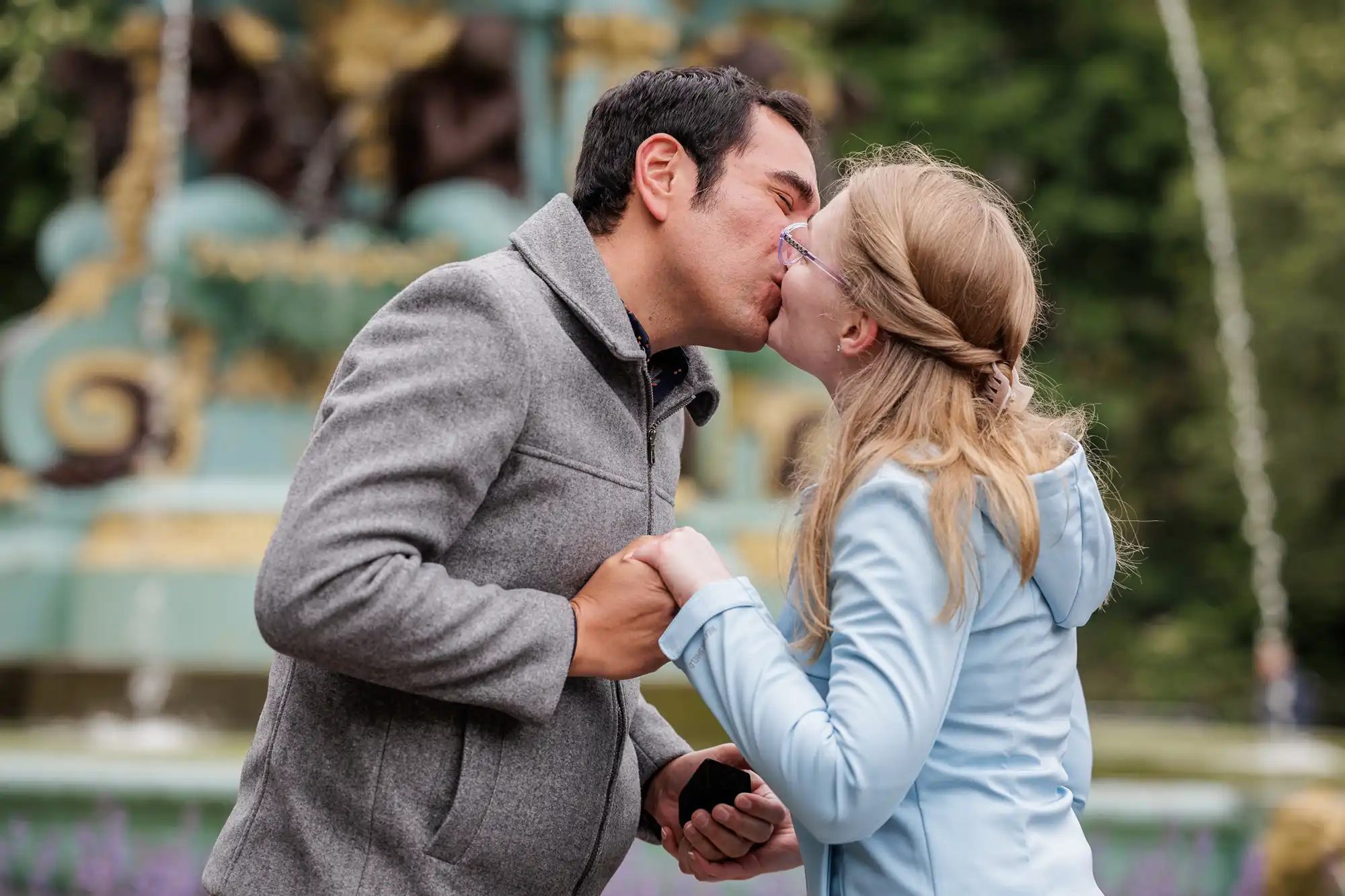 A man and woman kiss outdoors in front of a decorative fountain, holding hands. The man is holding a small black box, suggesting a marriage proposal.