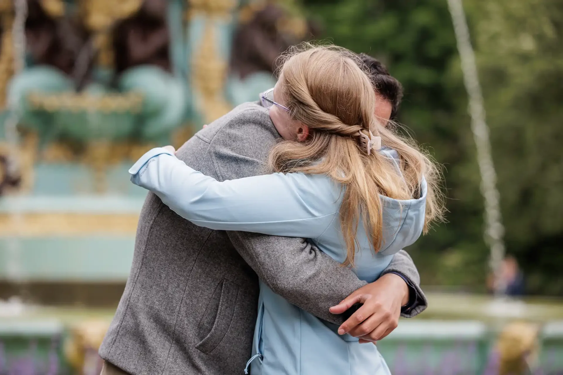 Two people embrace outdoors near a decorative fountain; one wears a light blue coat and the other a grey jacket.