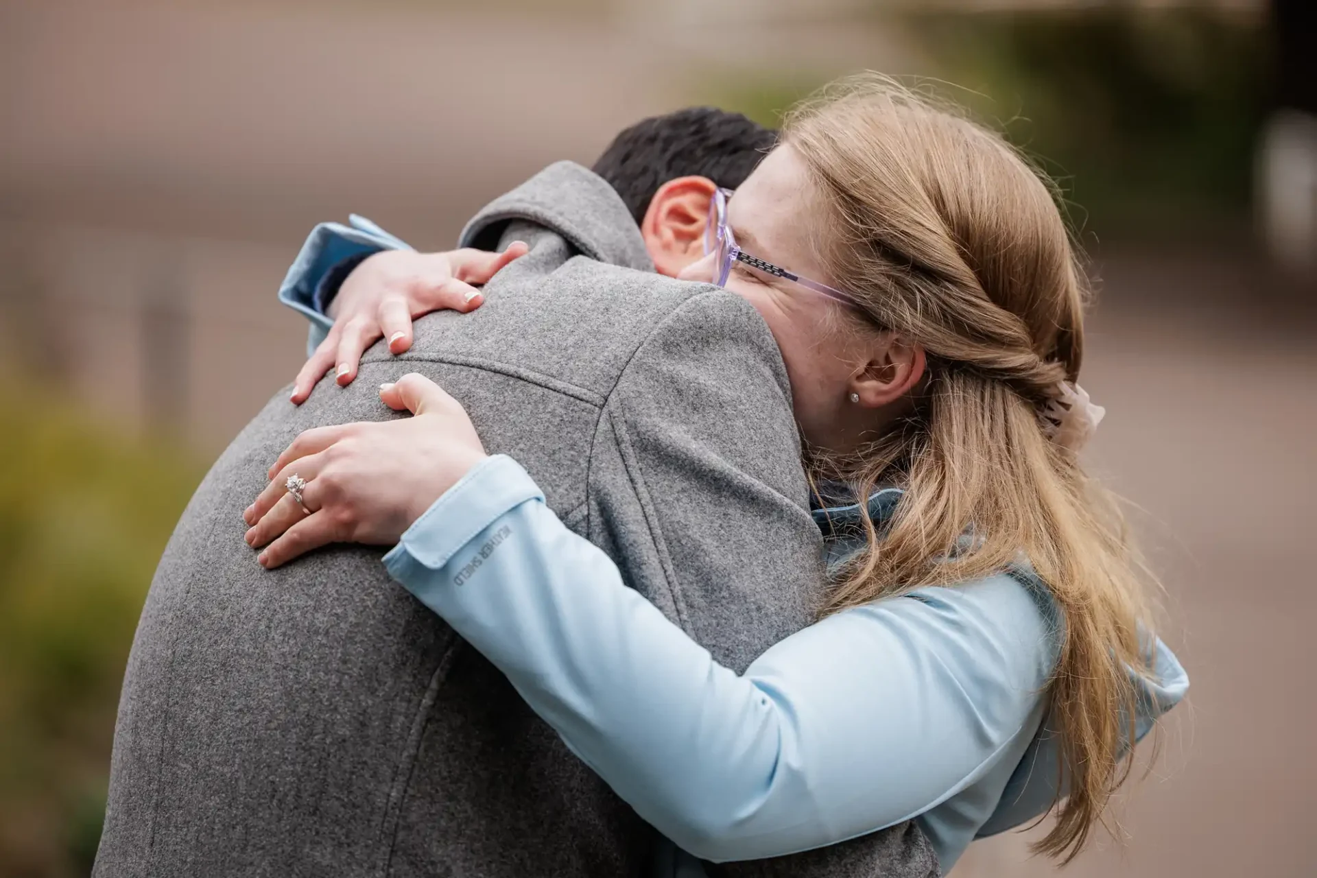 A woman wearing glasses and a light blue shirt hugs a man in a gray coat outdoors, both appearing happy and emotional.